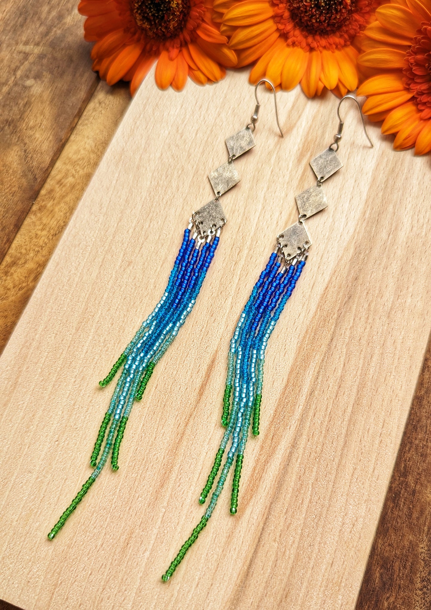 Moon & Milk beaded earrings with silver diamond connectors and long fringe in blue, teal, and green beads, styled with vibrant orange flowers.