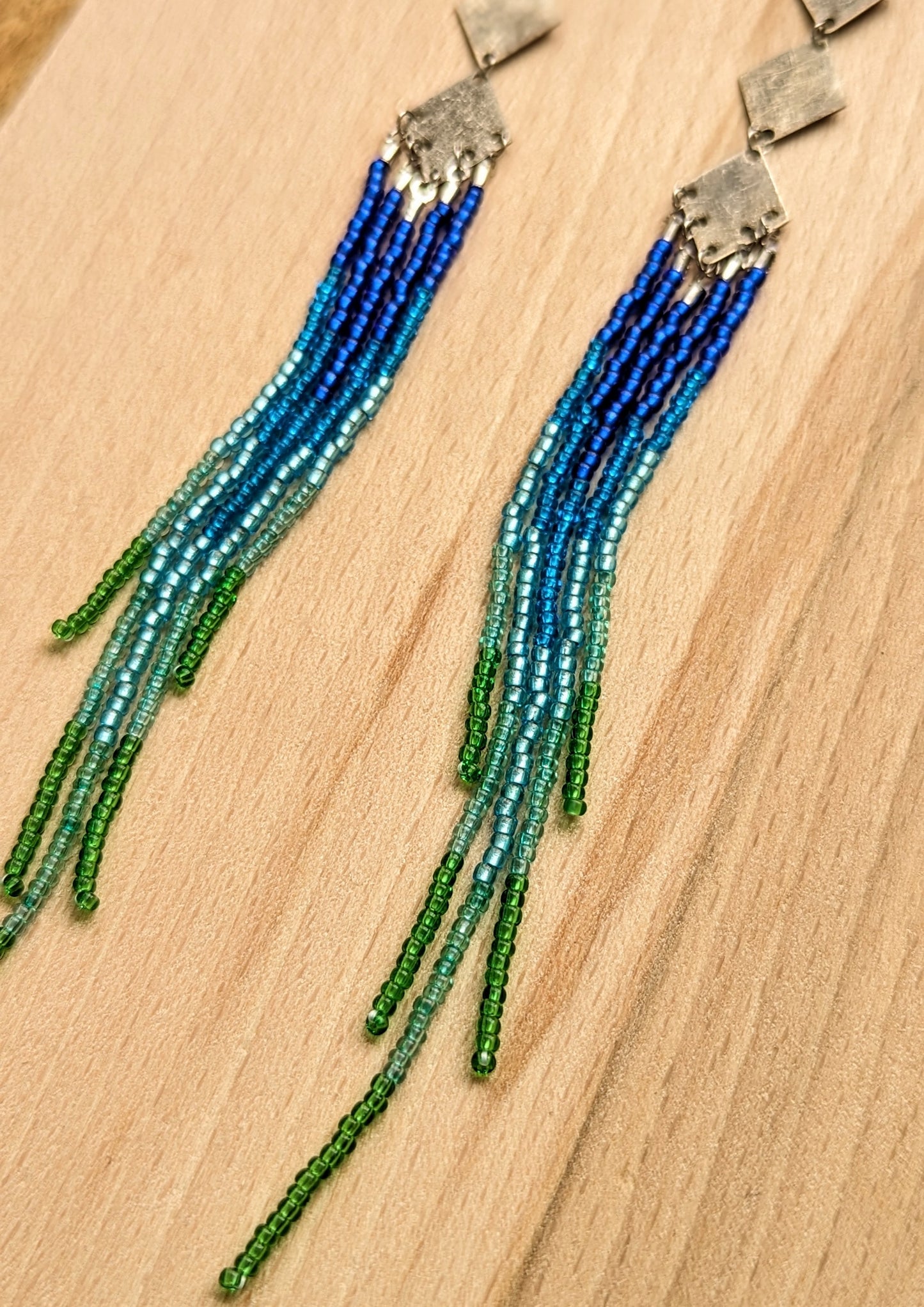 Moon & Milk beaded earrings with silver diamond connectors and long fringe in blue, teal, and green beads.
