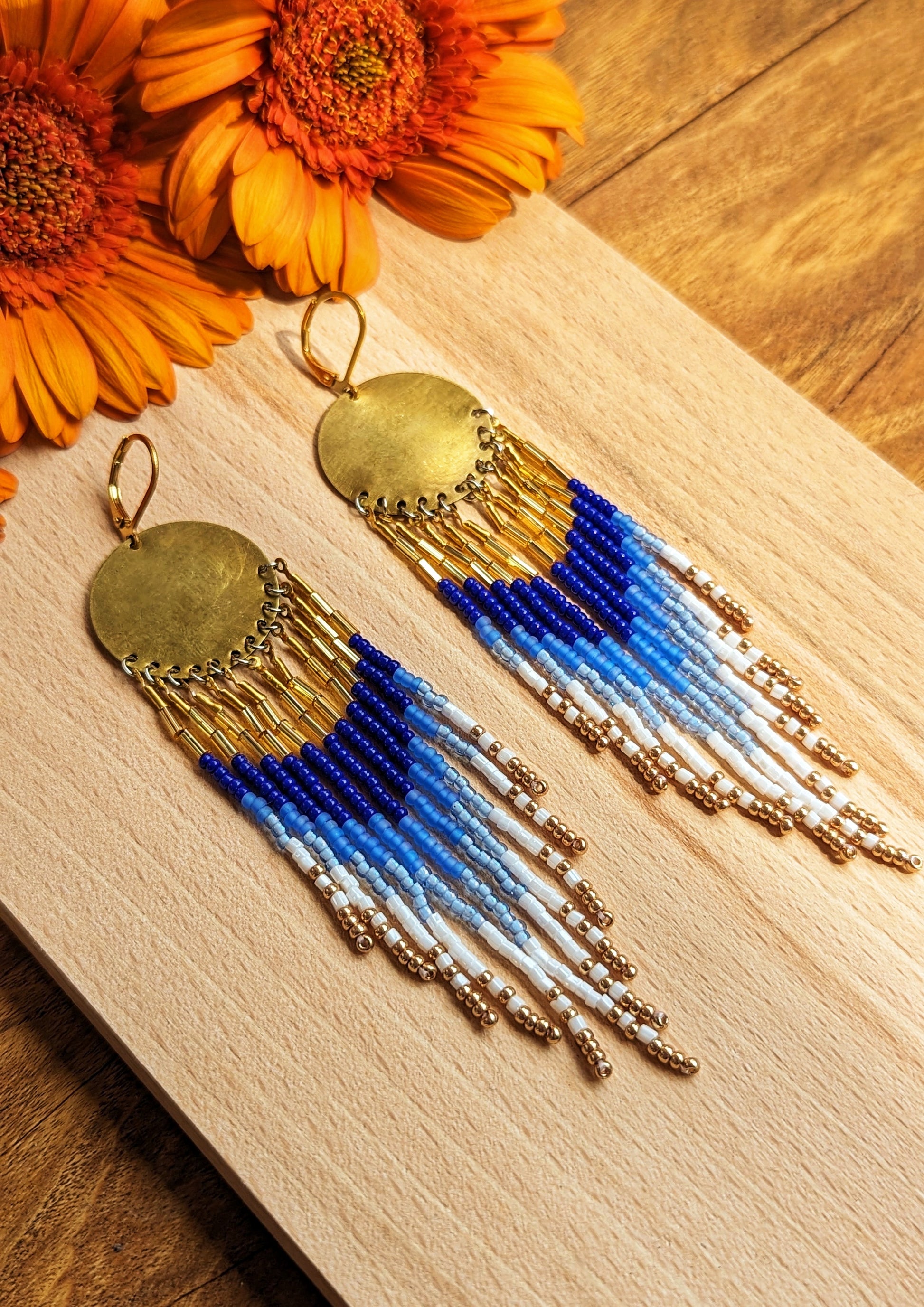 Moon & Milk hand-beaded earrings with cascading beaded fringes in cobalt blue, white, and gold tones, displayed on wood beside a bright orange flower.