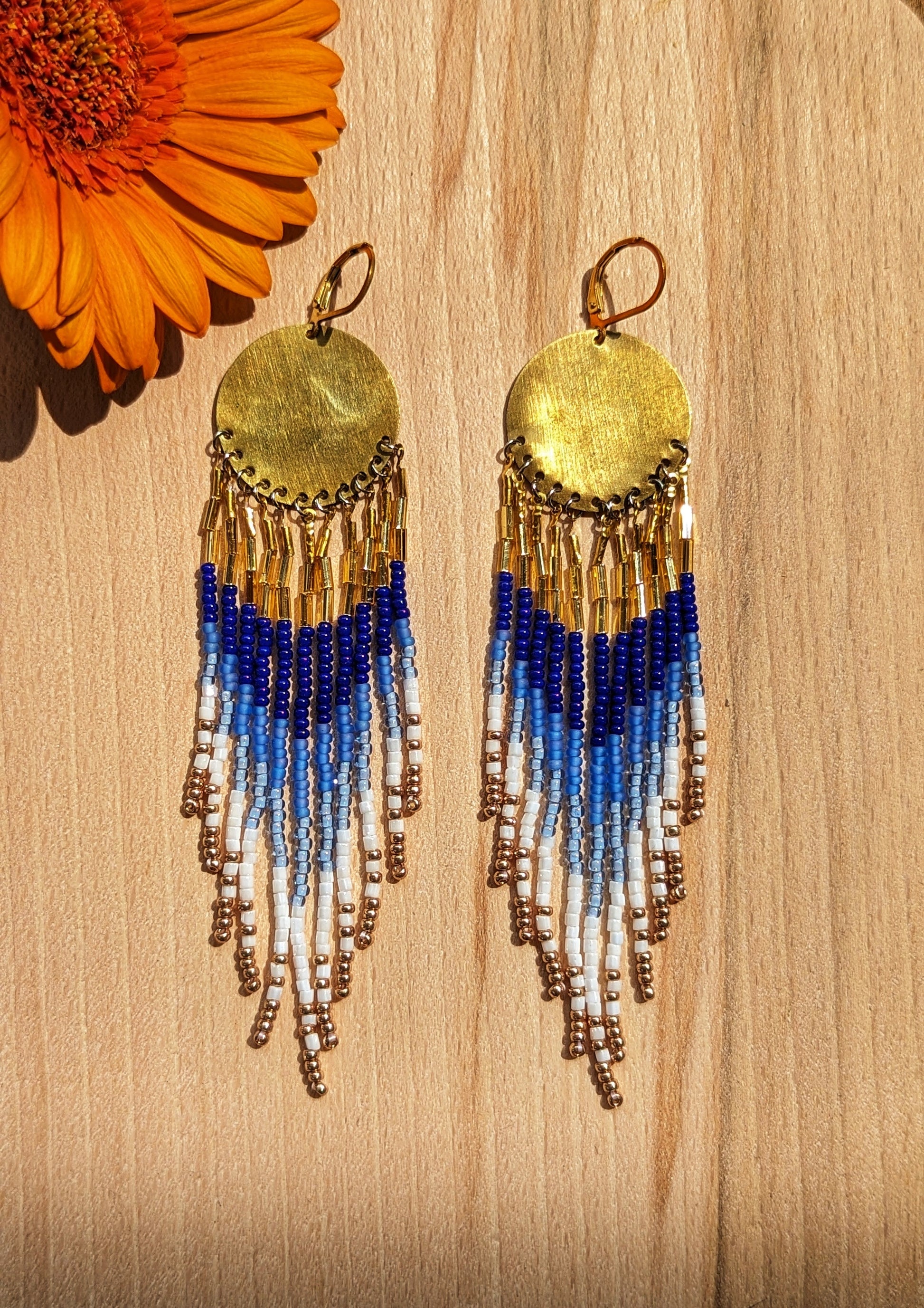 Moon & Milk hand-beaded earrings with cascading beaded fringes in cobalt blue, white, and gold tones, displayed on wood beside a bright orange flower.