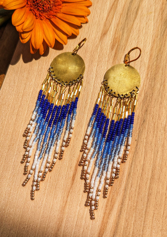 Moon & Milk hand-beaded earrings with cascading beaded fringes in cobalt blue, white, and gold tones, displayed on wood beside a bright orange flower.