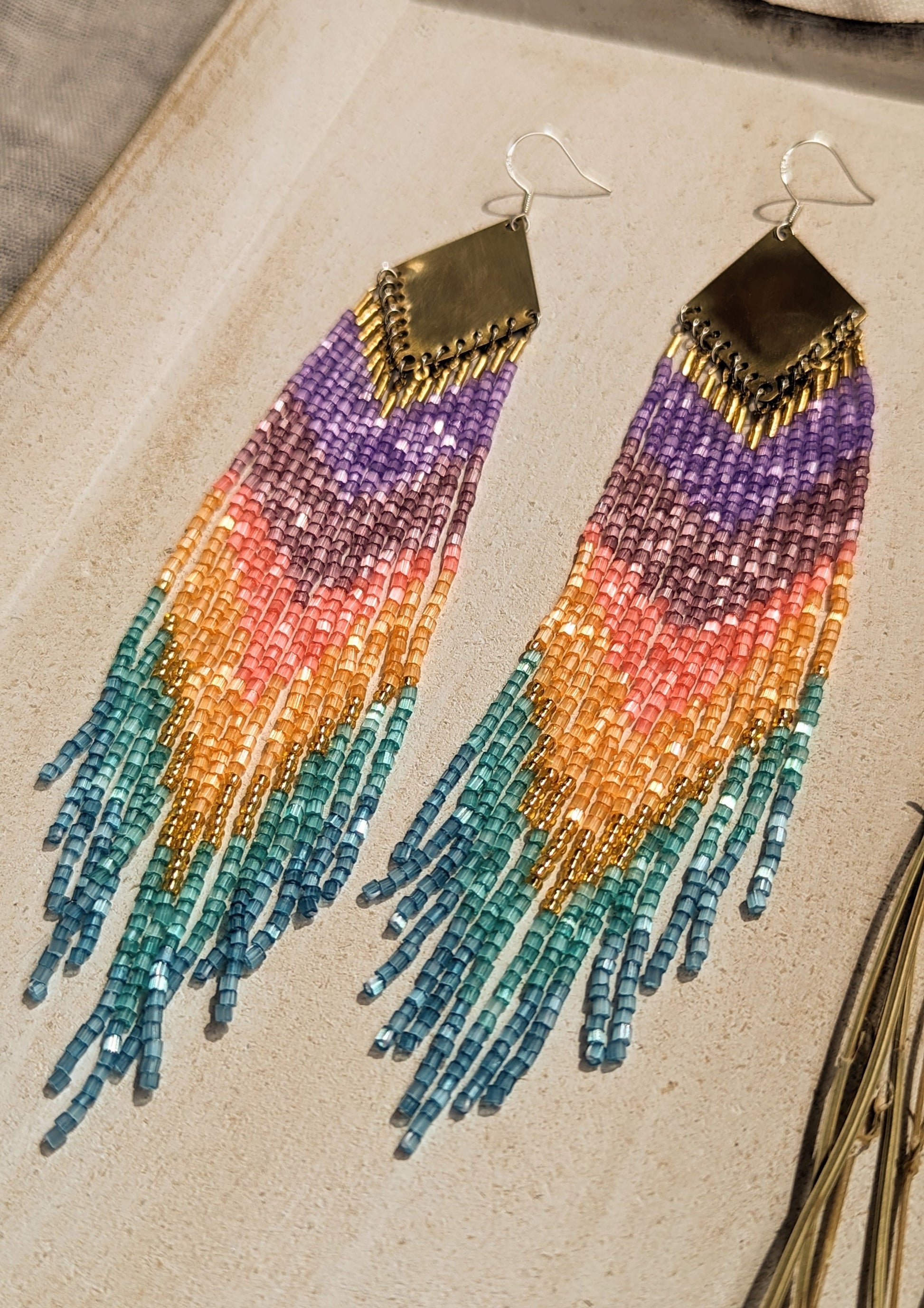 Moon & Milk rainbow ombré beaded earrings with purple, amethyst, coral, gold, and turquoise fringe hanging from brass diamond findings, displayed beside dried lavender.