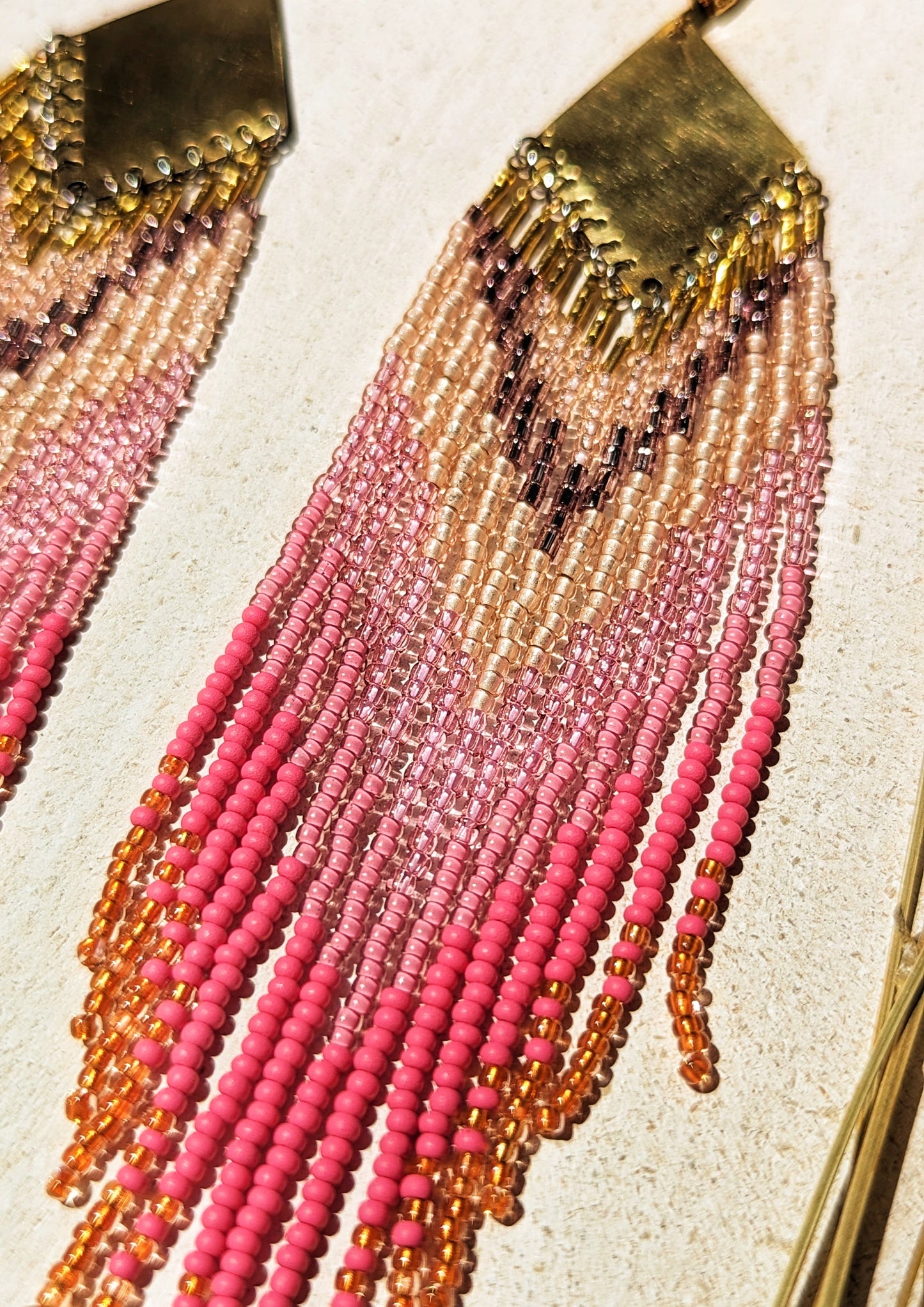 Moon & Milk - Handcrafted seed bead earrings with a vibrant gradient of pink, peach, and gold beads, elegantly hanging from a brass chevron pendant. Perfect boho-chic jewelry for adding a pop of color and style. Set against a white wooden tray.