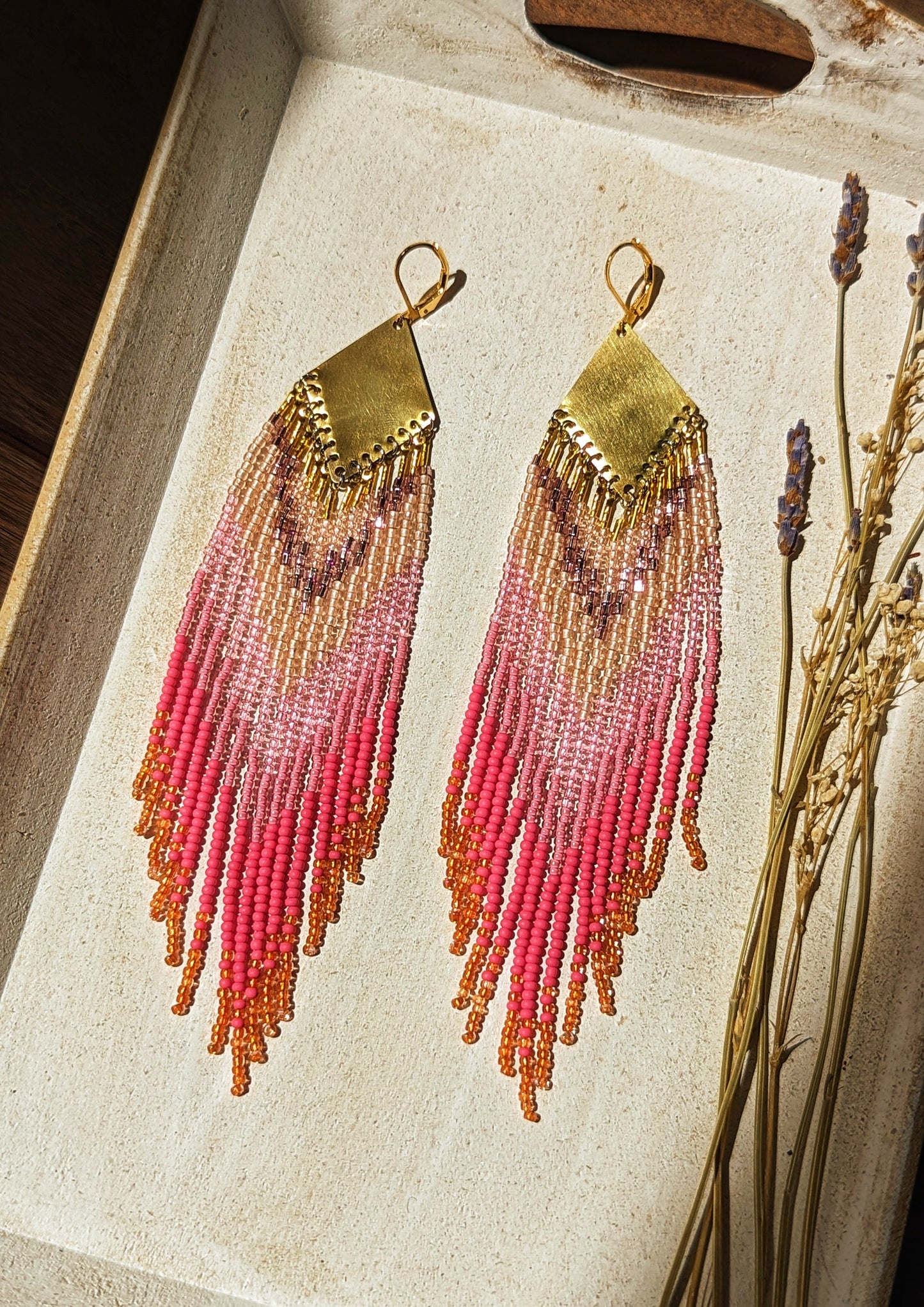 Moon & Milk - Handcrafted seed bead earrings with a vibrant gradient of pink, peach, and gold beads, elegantly hanging from a brass chevron pendant. Perfect boho-chic jewelry for adding a pop of color and style. Set against a white wooden tray.