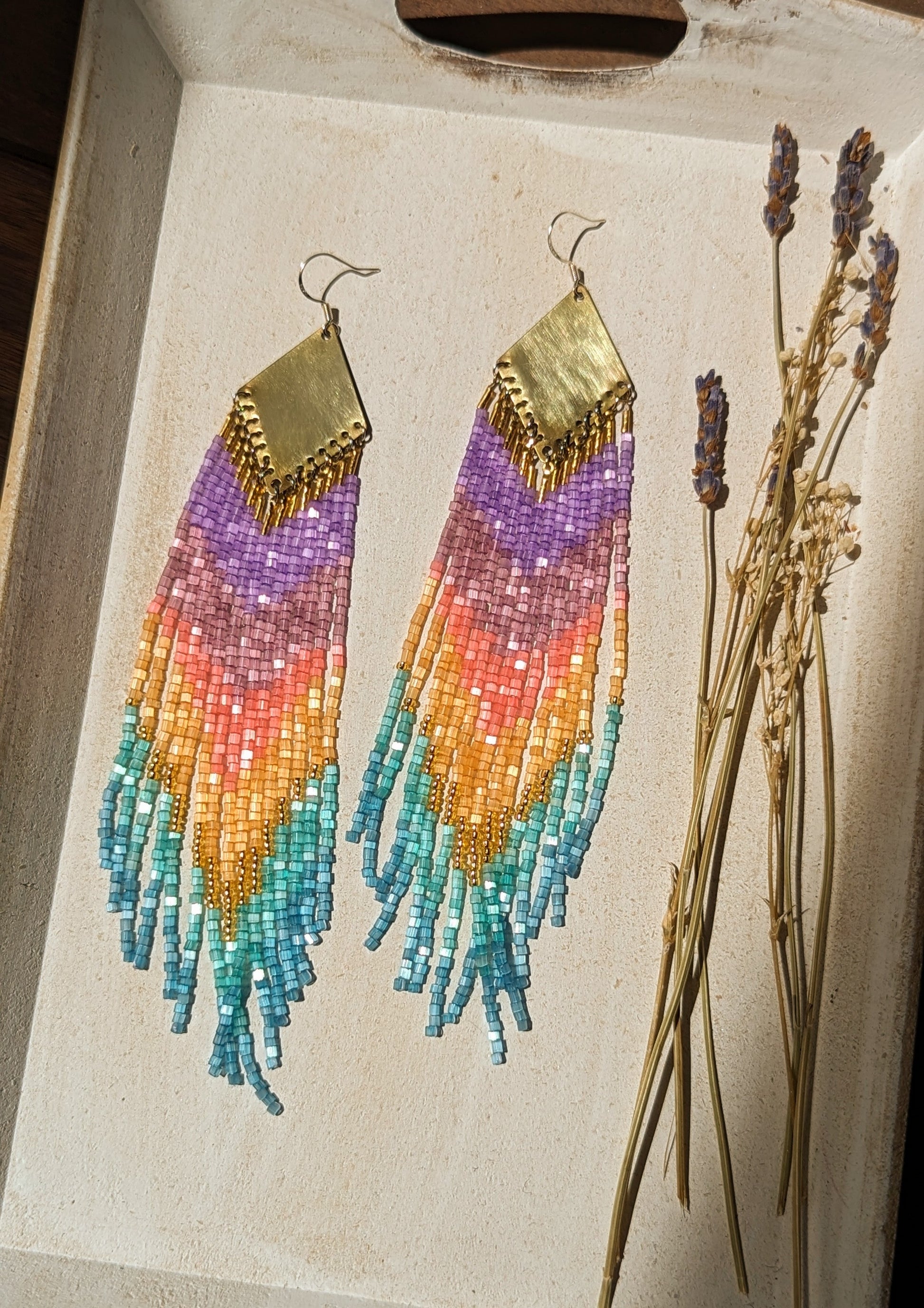 Moon & Milk rainbow ombré beaded earrings with purple, amethyst, coral, gold, and turquoise fringe hanging from brass diamond findings, displayed beside dried lavender.