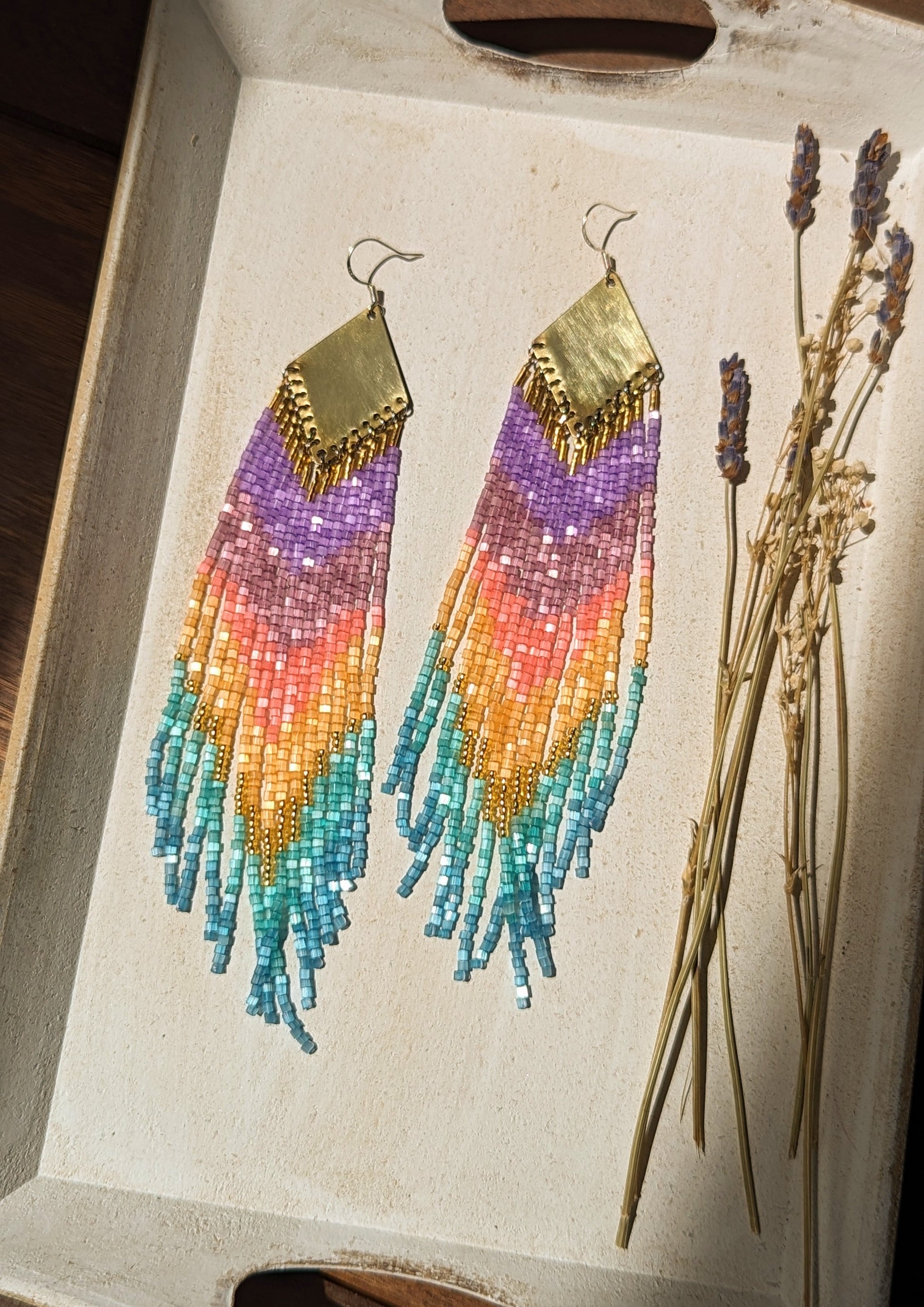 Moon & Milk rainbow ombré beaded earrings with purple, amethyst, coral, gold, and turquoise fringe hanging from brass diamond findings, displayed beside dried lavender.