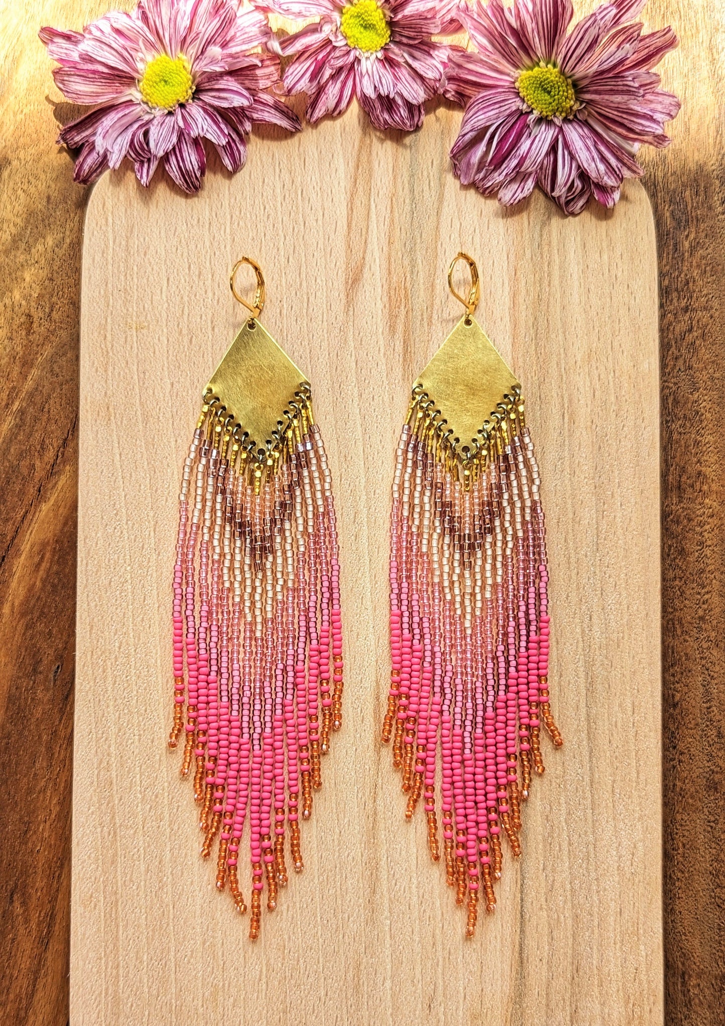 Moon & Milk - Handcrafted seed bead earrings with a vibrant gradient of pink, peach, and gold beads, elegantly hanging from a brass chevron pendant. Perfect boho-chic jewelry for adding a pop of color and style. Set against a wooden background with delicate flowers.