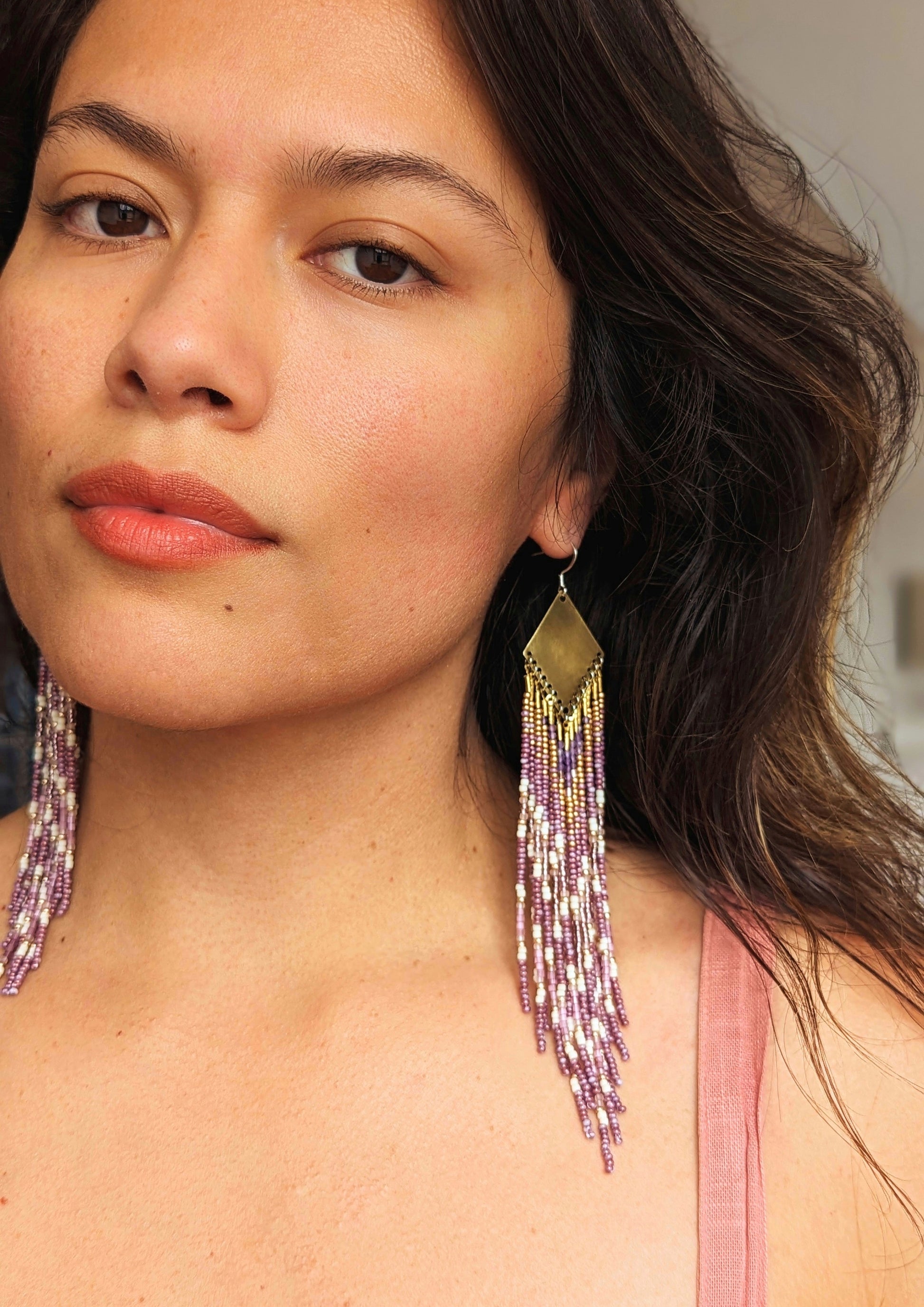 Moon & Milk long beaded earrings in gold, lavender, and cream tones, handcrafted with glass beads and brass worn by a woman for a soft, radiant native-inspired fringe design.