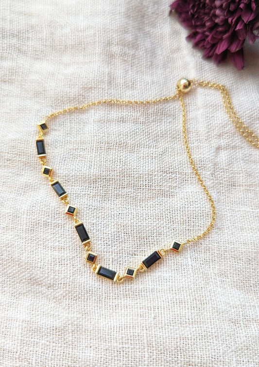 Gold chain bracelet with alternating black rectangular and black crystals displayed flat on textured fabric with a gold slider clasp.