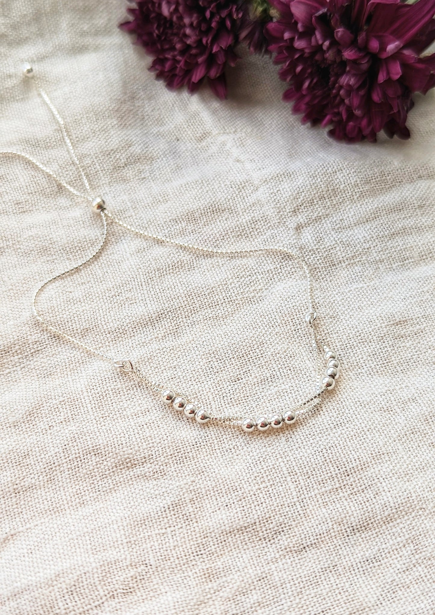 Delicate silver adjustable bracelet with a minimalist design featuring polished beads and a sliding clasp, displayed on a linen white cloth with floral accents.