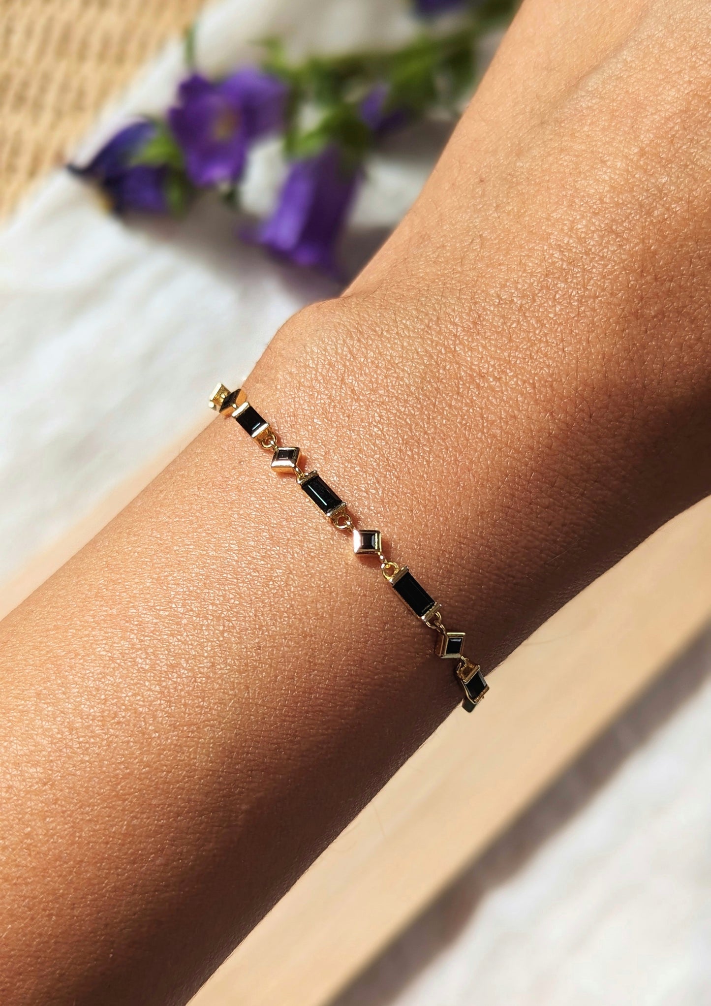Minimalist black and gold bracelet with geometric stones worn on wrist, shown in natural sunlight with blurred purple flowers in the background.