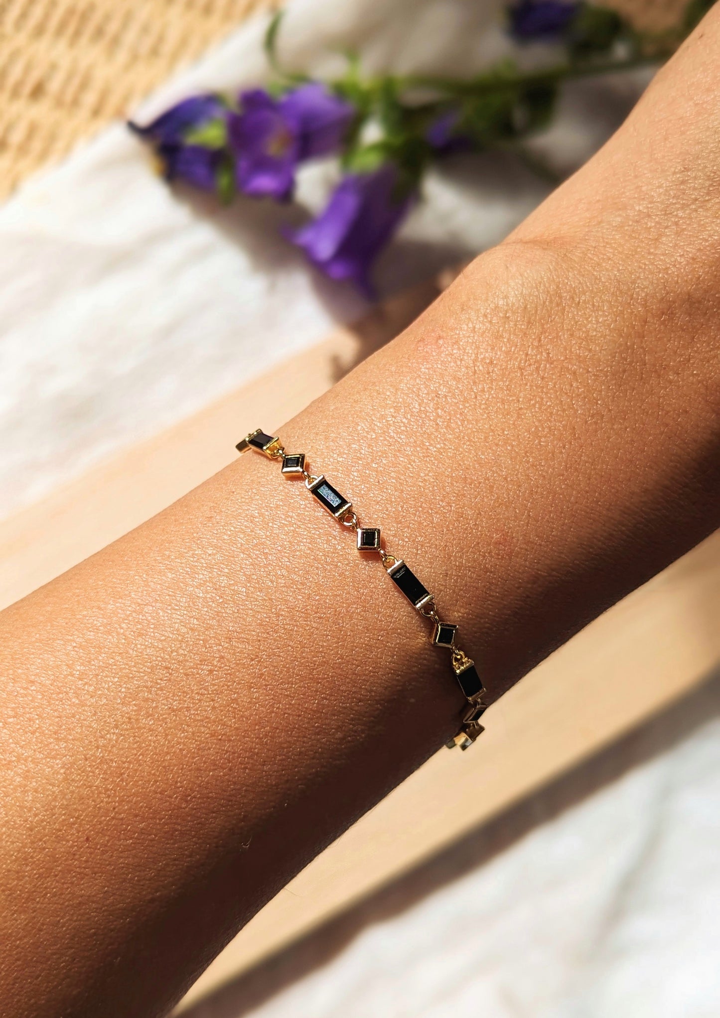 Minimalist black and gold bracelet with geometric stones worn on wrist, shown in natural sunlight with blurred purple flowers in the background.