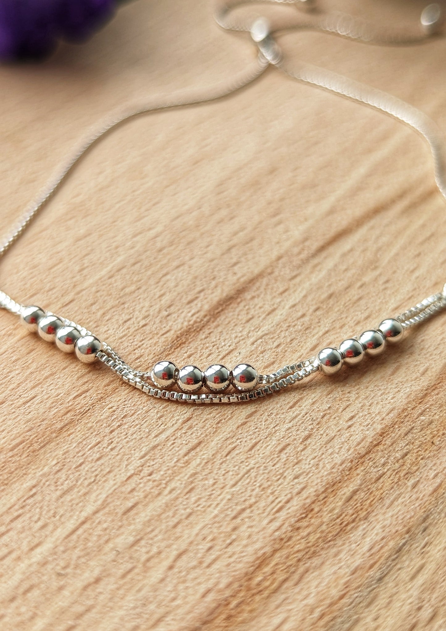 Delicate silver adjustable bracelet with a minimalist design featuring polished beads and a sliding clasp, displayed on a wooden surface with floral accents.