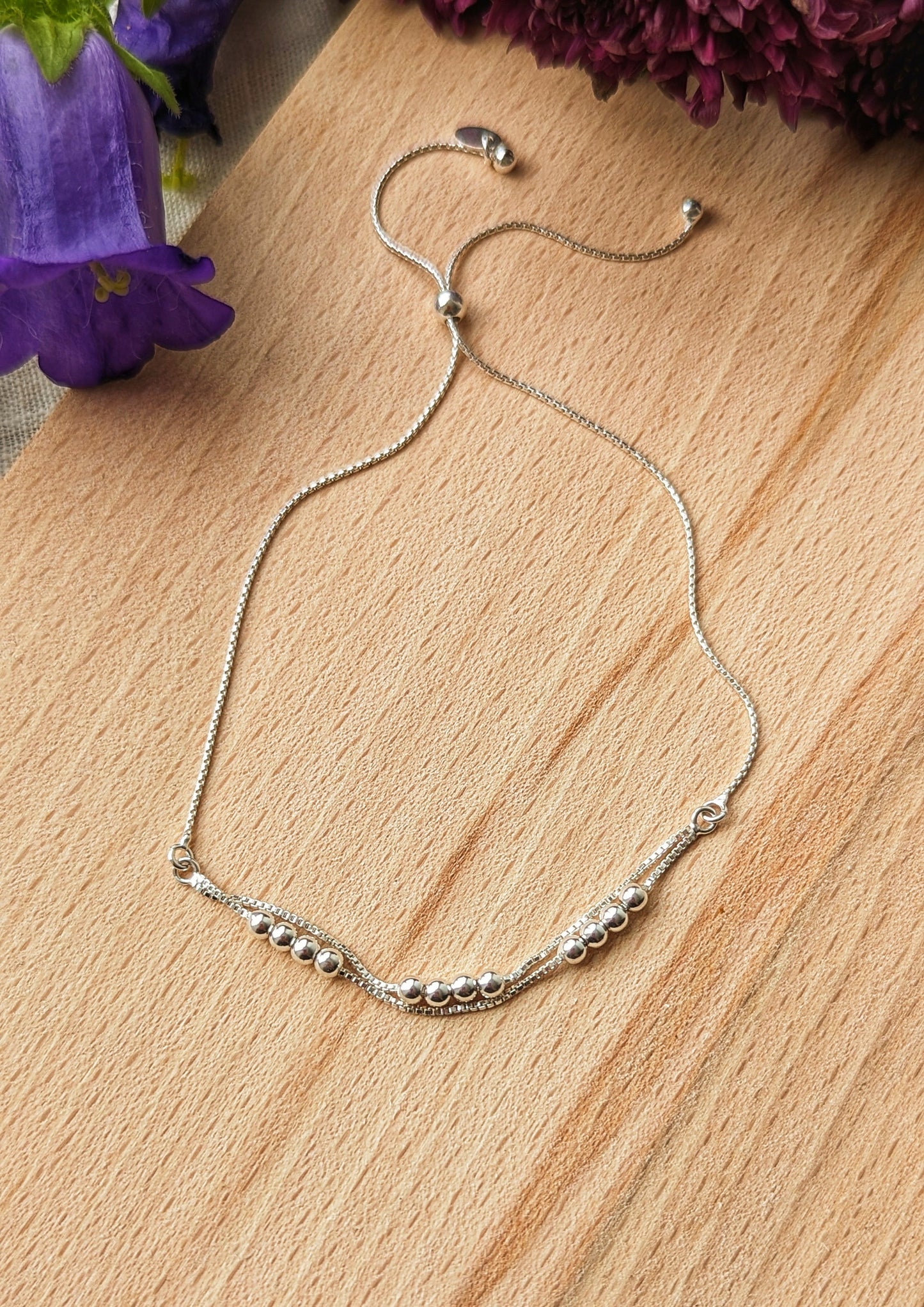 Delicate silver adjustable bracelet with a minimalist design featuring polished beads and a sliding clasp, displayed on a wooden surface with floral accents.