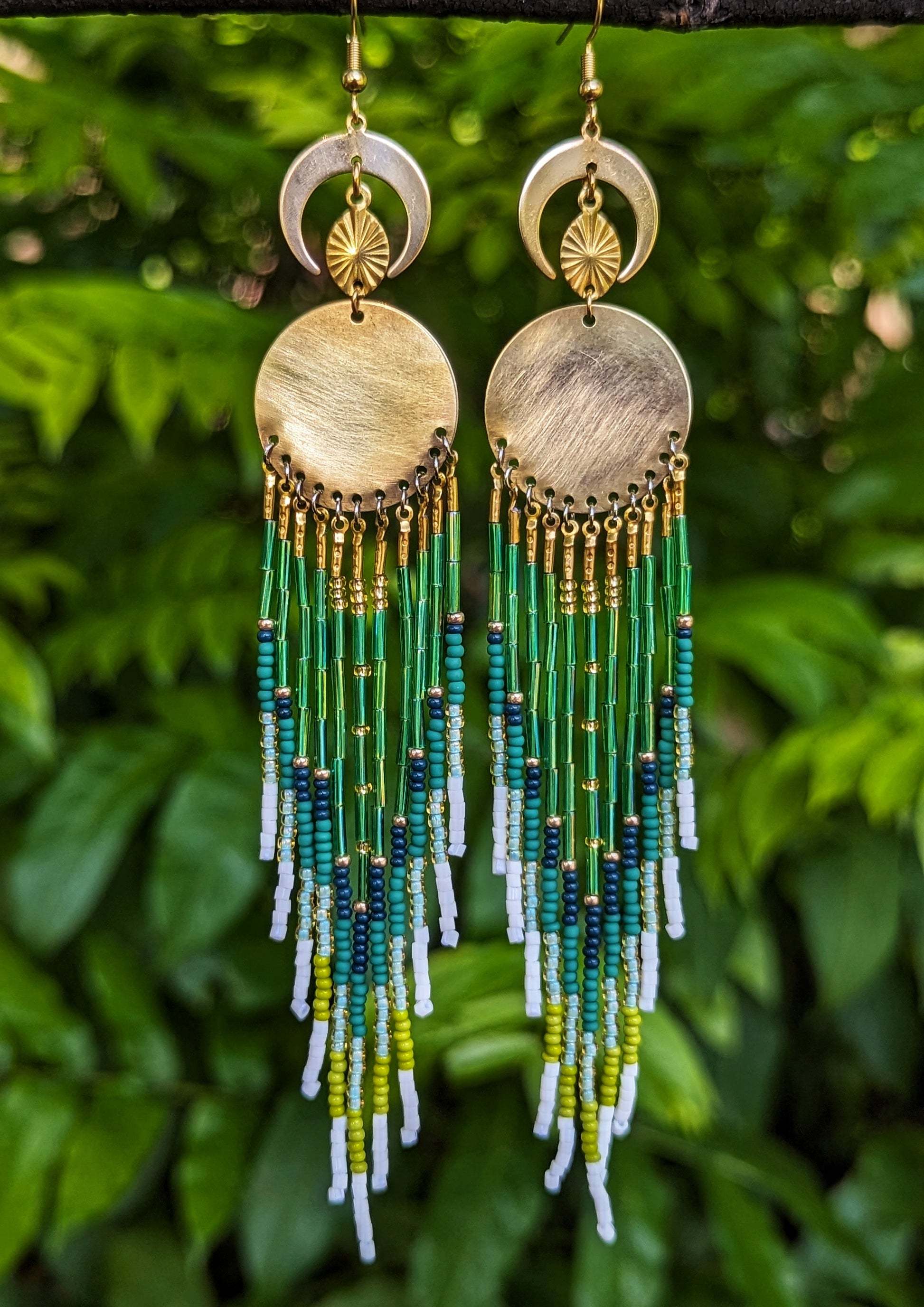 Moon & Milk handcrafted long native earrings with green, white, and gold beadwork, brass discs, and crescent moons, hanging from a branch.