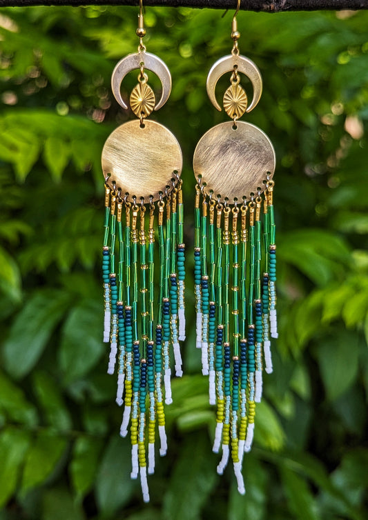 Moon & Milk handcrafted long native earrings with green, white, and gold beadwork, brass discs, and crescent moons, hanging from a branch.