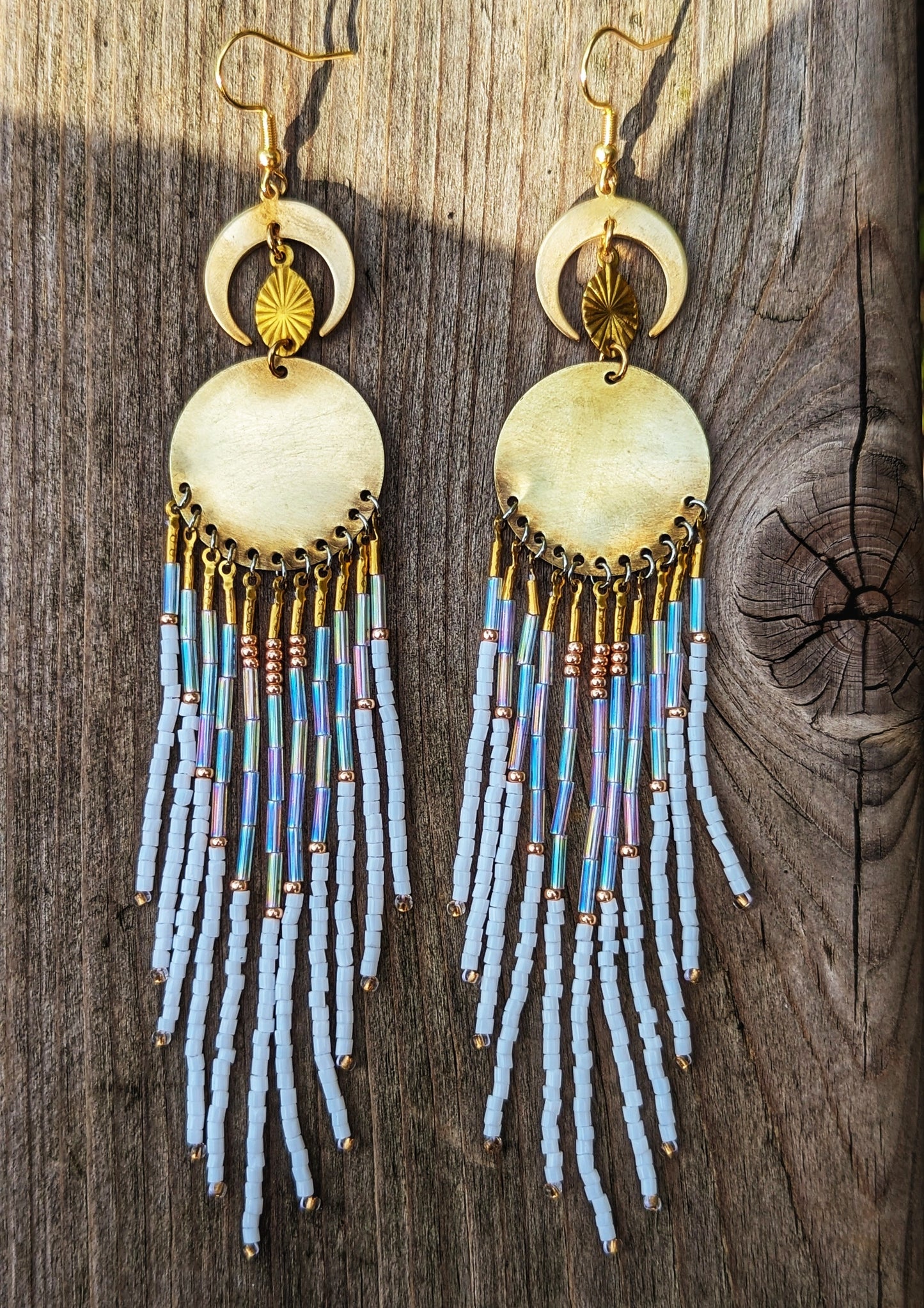 Moon & Milk beaded earrings with gold crescent moons and discs, featuring cascading white, iridescent, and copper-toned fringes on a wooden background.