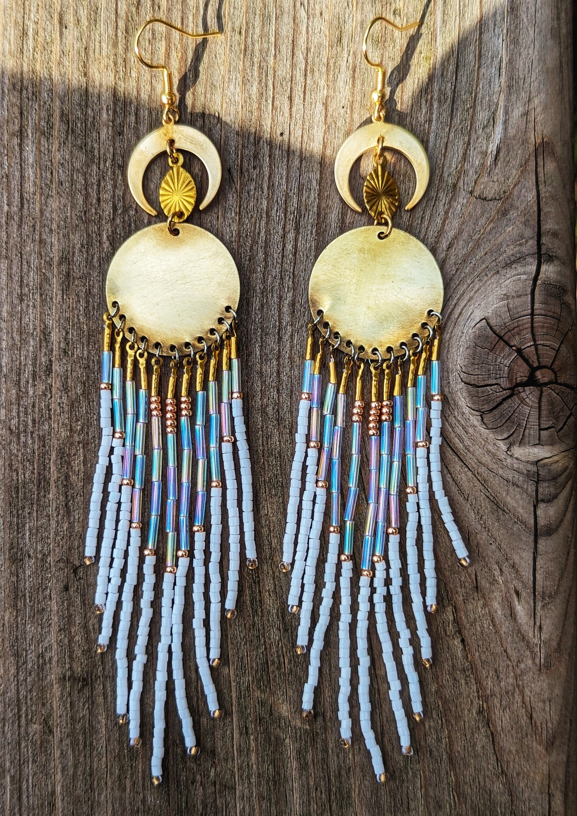 Moon & Milk beaded earrings with gold crescent moons and discs, featuring cascading white, iridescent, and copper-toned fringes on a wooden background.