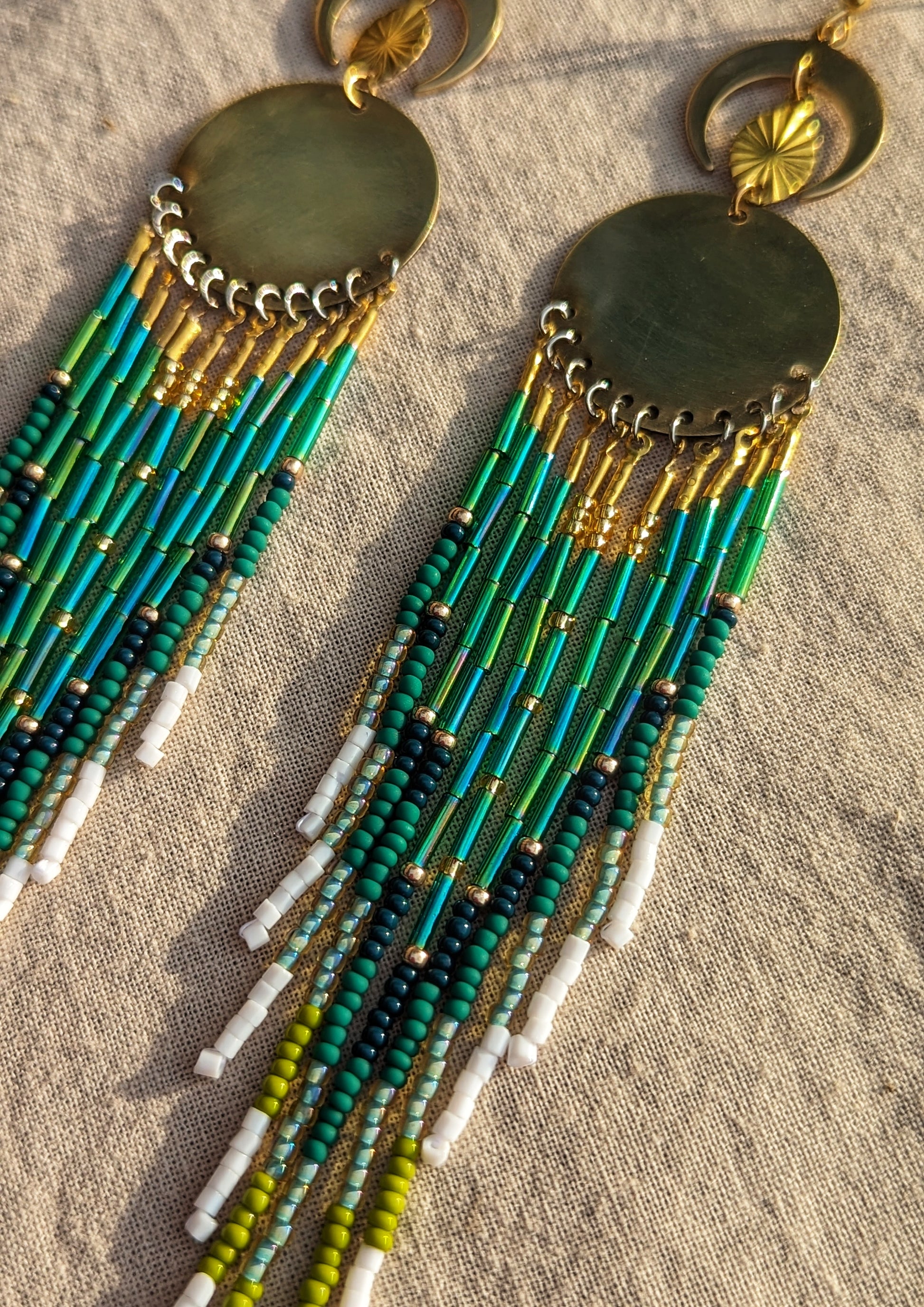 Moon & Milk handcrafted long native earrings with green, white, and gold beadwork, brass discs, and crescent moons, laid on a tan fabric background.