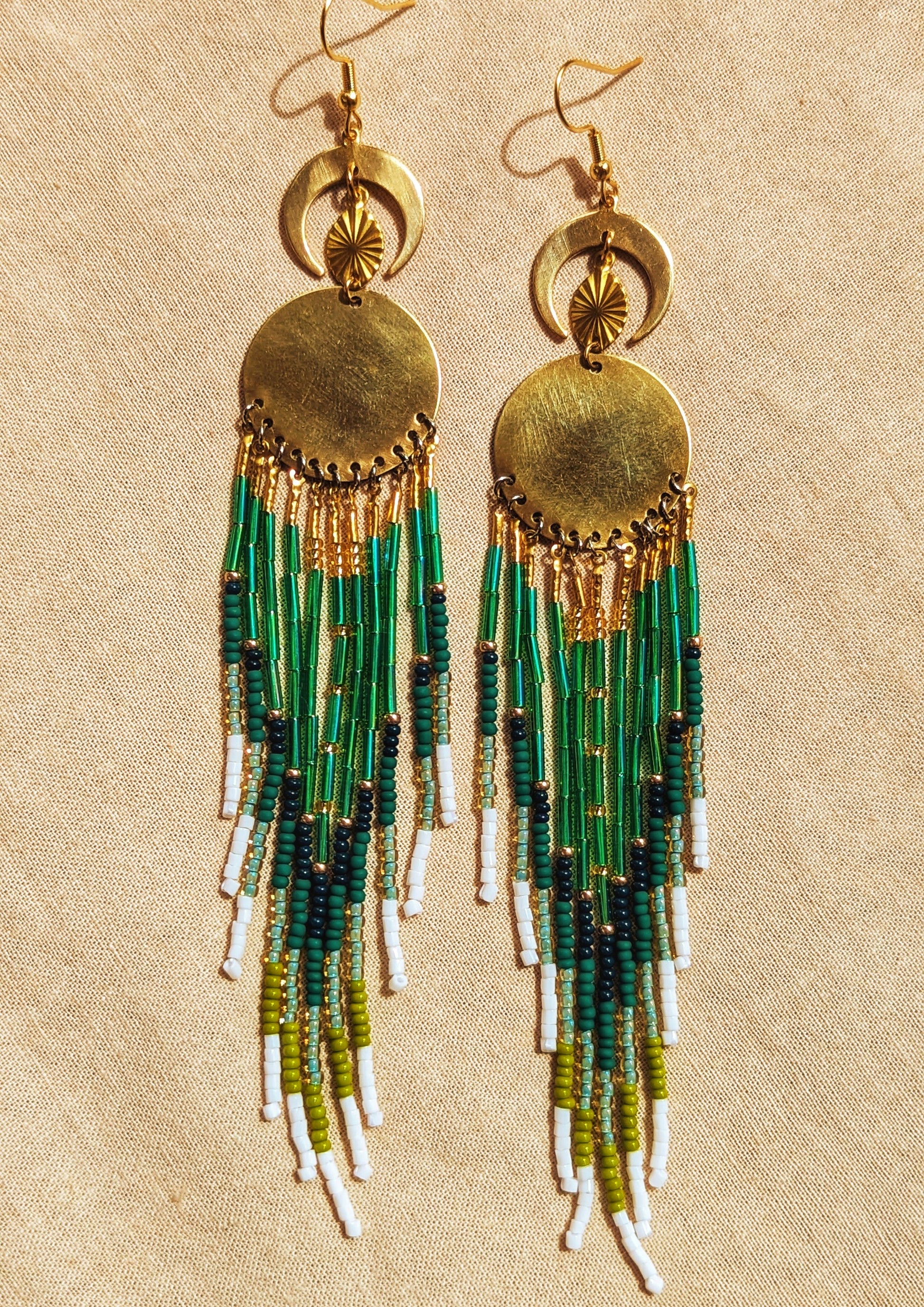 Moon & Milk handcrafted long native earrings with green, white, and gold beadwork, brass discs, and crescent moons, laid on a tan fabric background.
