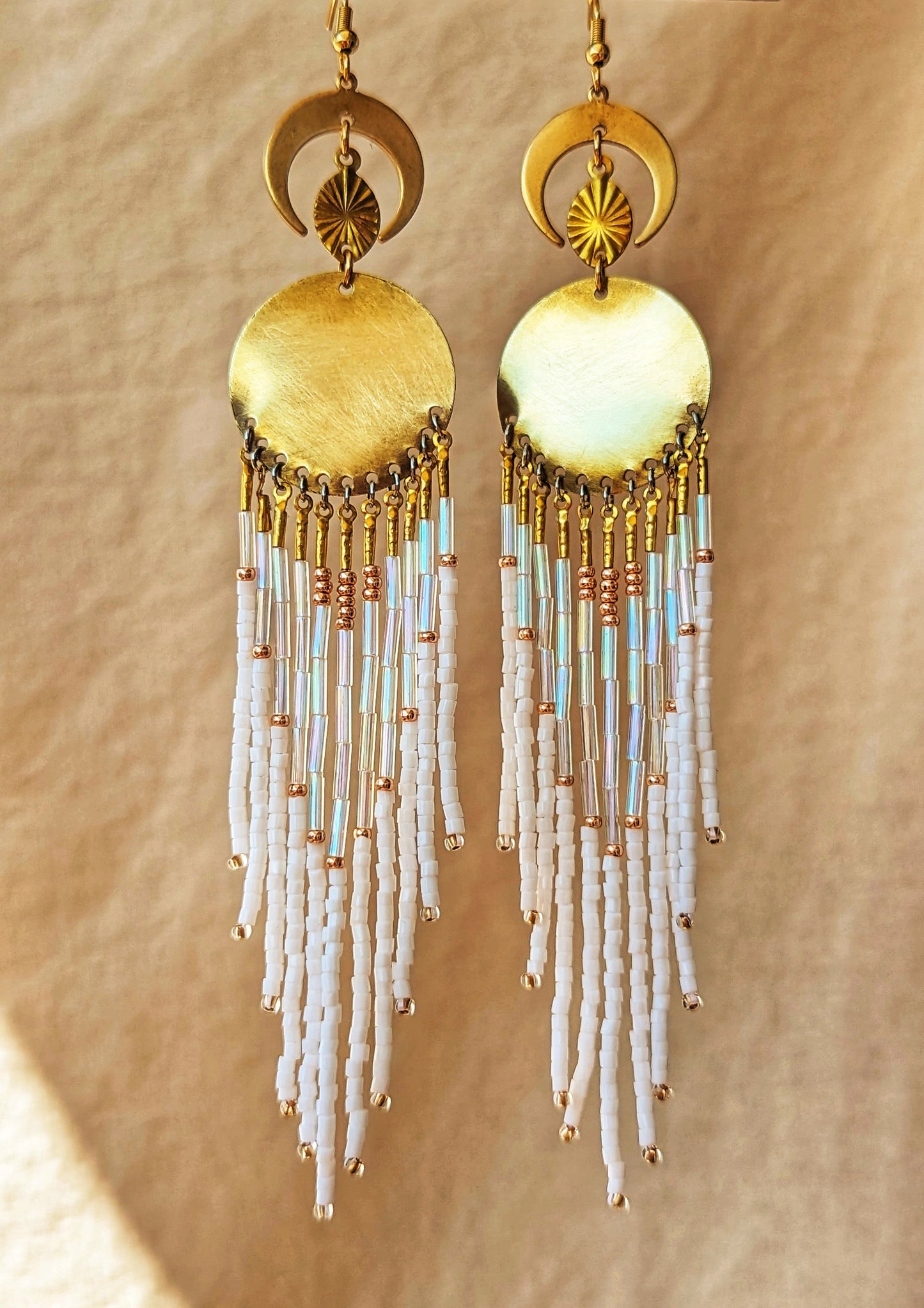 Moon & Milk beaded earrings with gold crescent moons and discs, featuring cascading white, iridescent, and copper-toned fringe son a warm beige fabric backdrop.