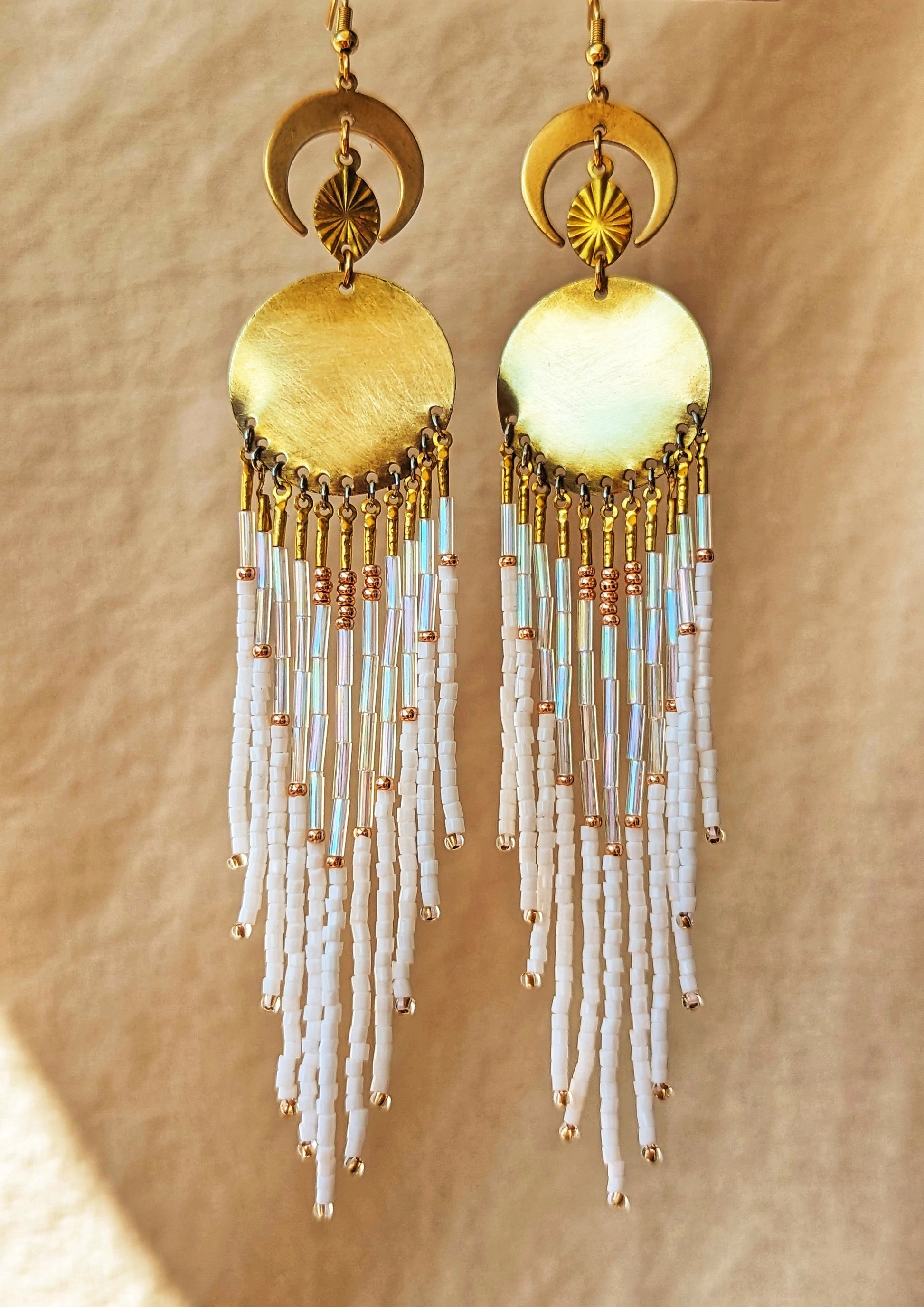 Moon & Milk beaded earrings with gold crescent moons and discs, featuring cascading white, iridescent, and copper-toned fringe son a warm beige fabric backdrop.