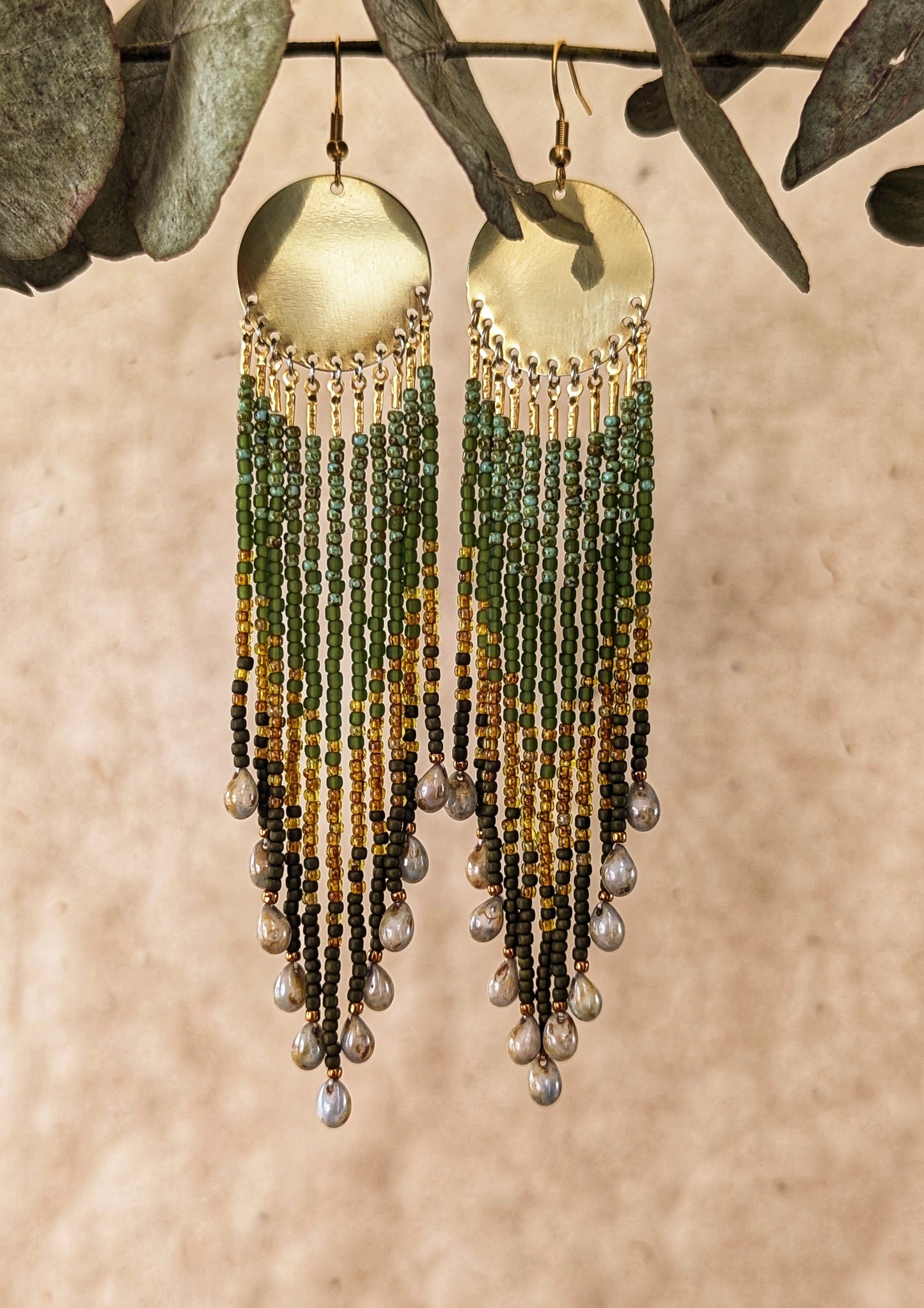 Boho beaded earrings made with brass disks, olive green seed beads, gold accents, and dangling glass teardrops on a neutral backdrop.