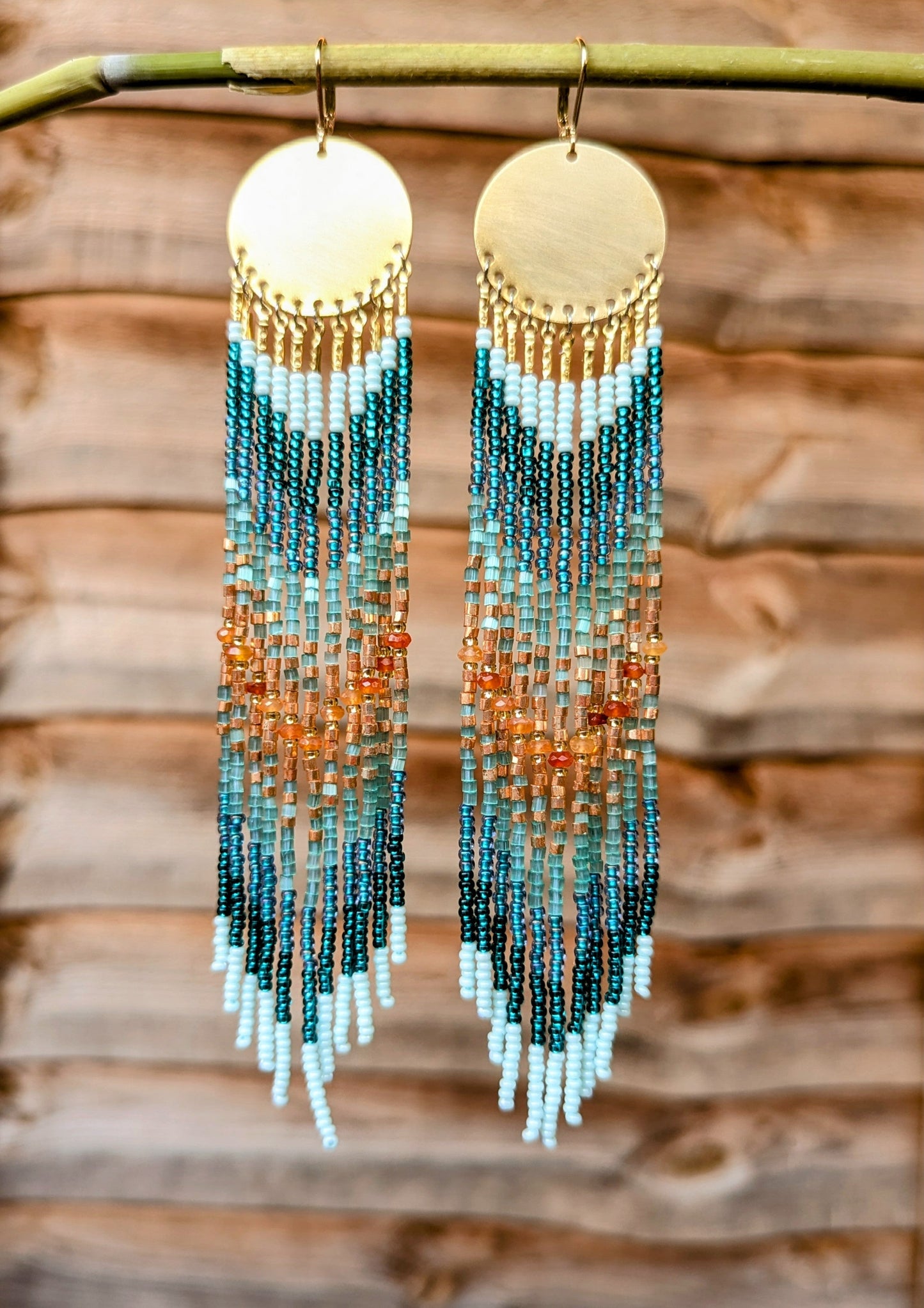 Hand beaded earrings with gradient colors hanging on a branch against a wooden background