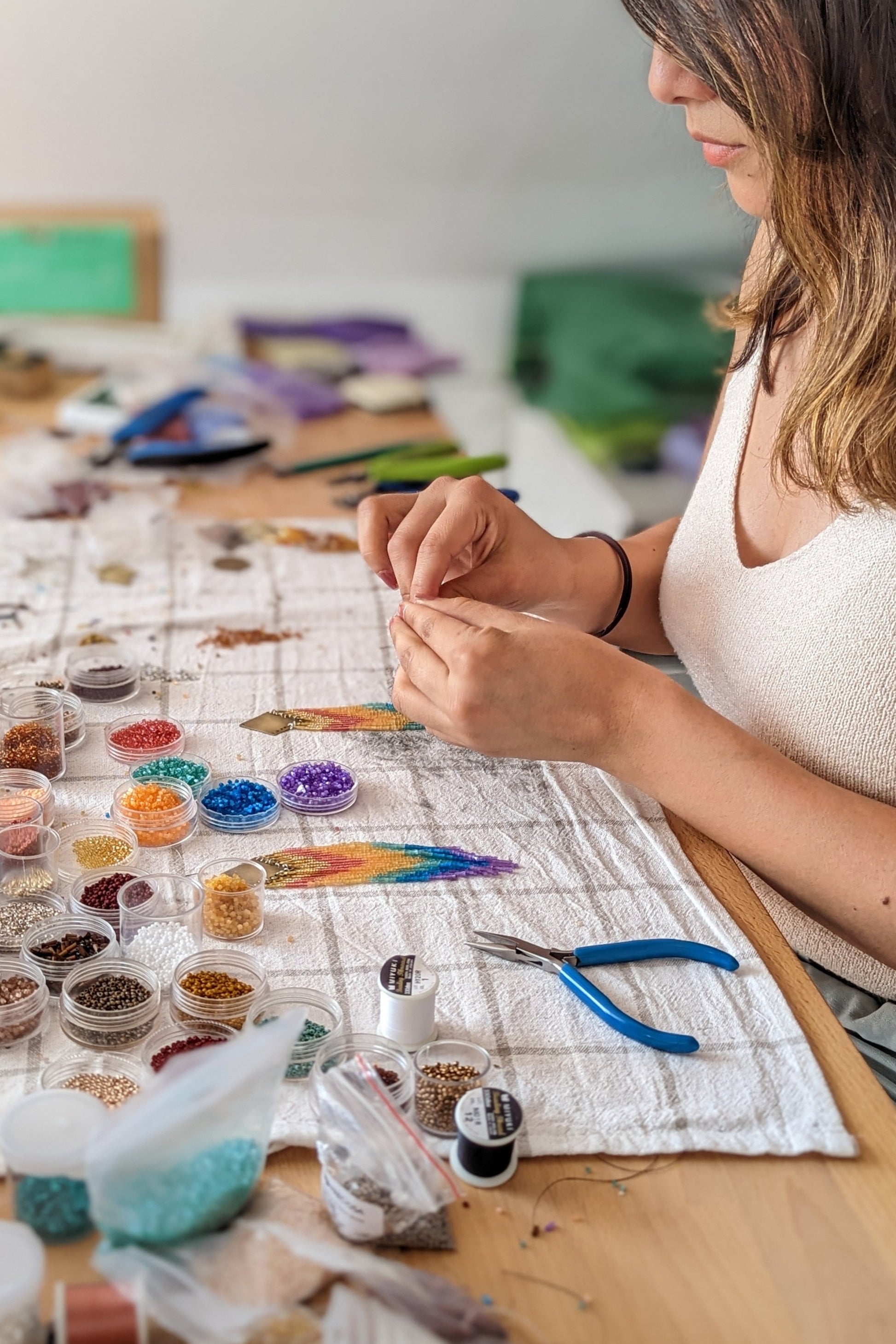 A woman handcrafting beaded earrings at a workspace covered in colorful beads, pliers, and thread—capturing the artistry behind Moon & Milk jewelry.