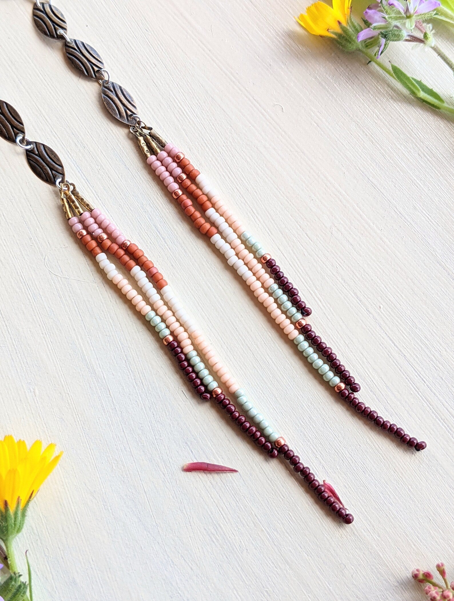 Moon & Milk handcrafted seed bead earrings with oval accents in blush pink, orange, cream, rust, mint, and burgundy, displayed with wildflowers on linen.