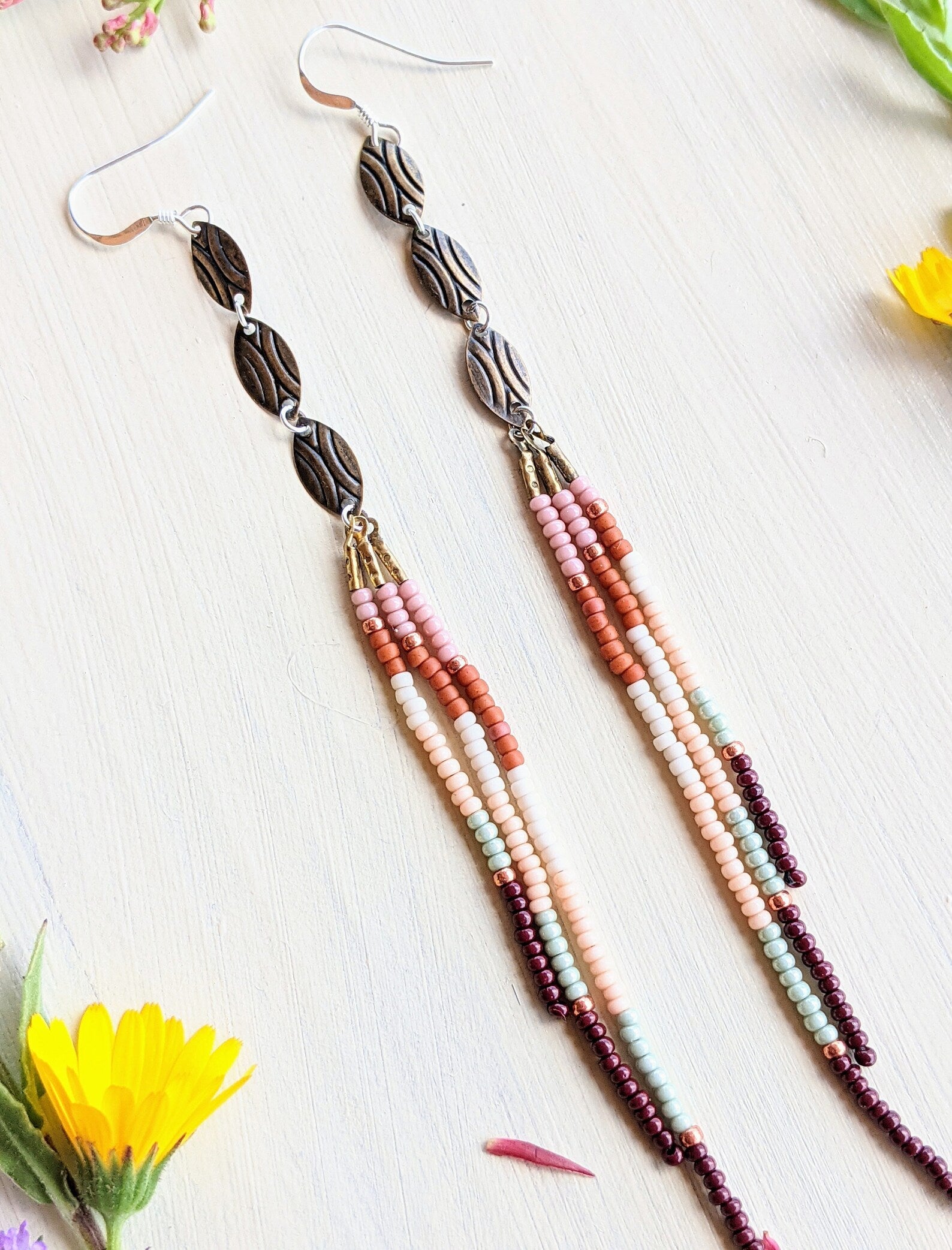 Moon & Milk handcrafted seed bead earrings with oval accents in blush pink, orange, cream, rust, mint, and burgundy, displayed with wildflowers on linen.