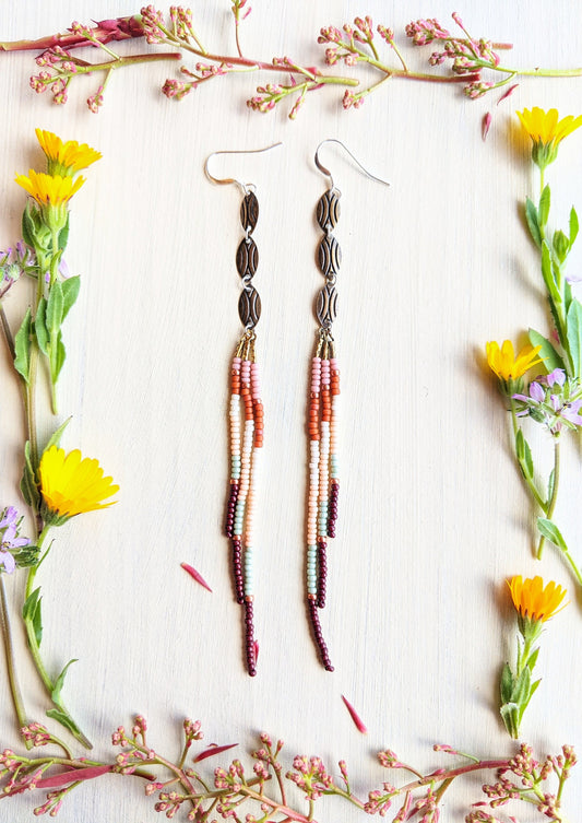 Moon & Milk handcrafted seed bead earrings with oval accents in blush pink, orange, cream, rust, mint, and burgundy, displayed with wildflowers on linen.