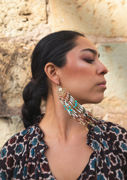 Woman wearing large, intricate beaded earrings against a stone wall background.