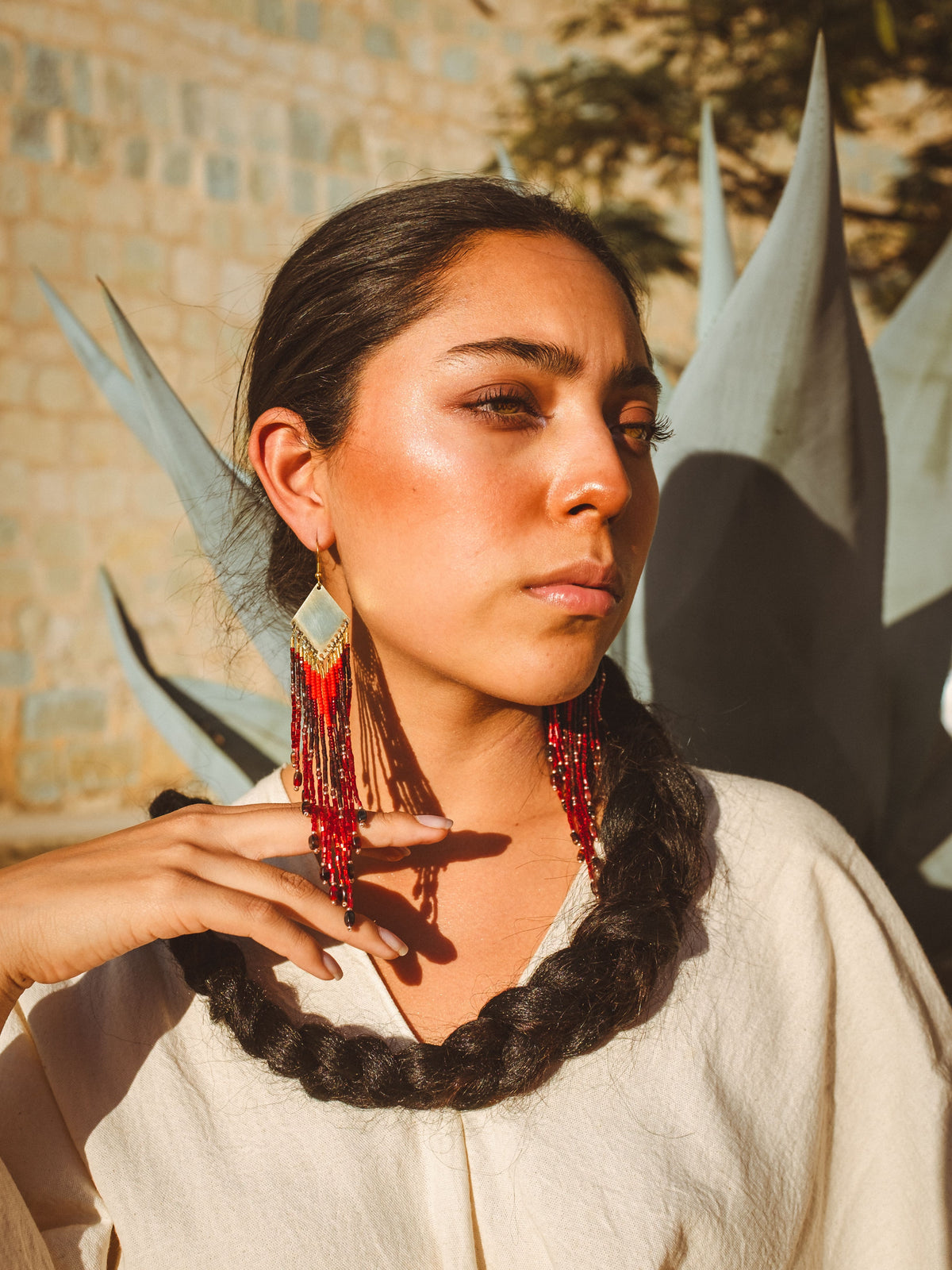 Woman with braided hair and boho red earrings in front of a textured wall and agave plant.