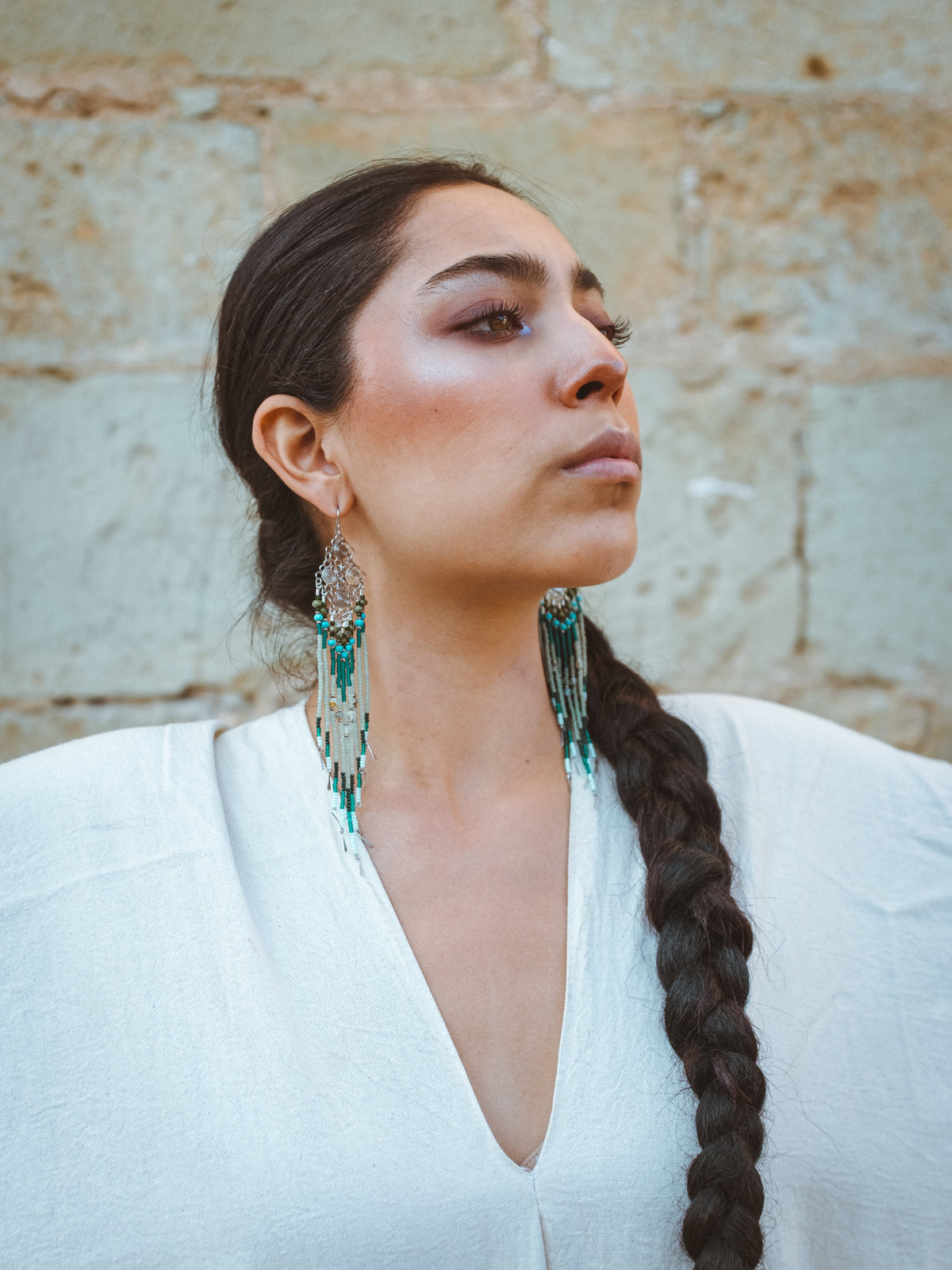 Woman with braided hair wearing artisanal beaded earrings and a white top against a textured wall.