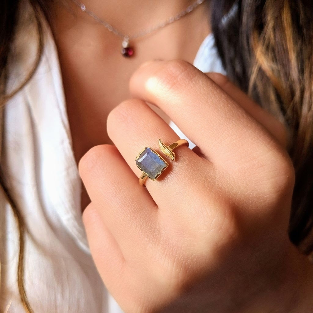 A gold-plated ring with a rectangular labradorite stone and a sculpted branch detail is shown worn on a hand.