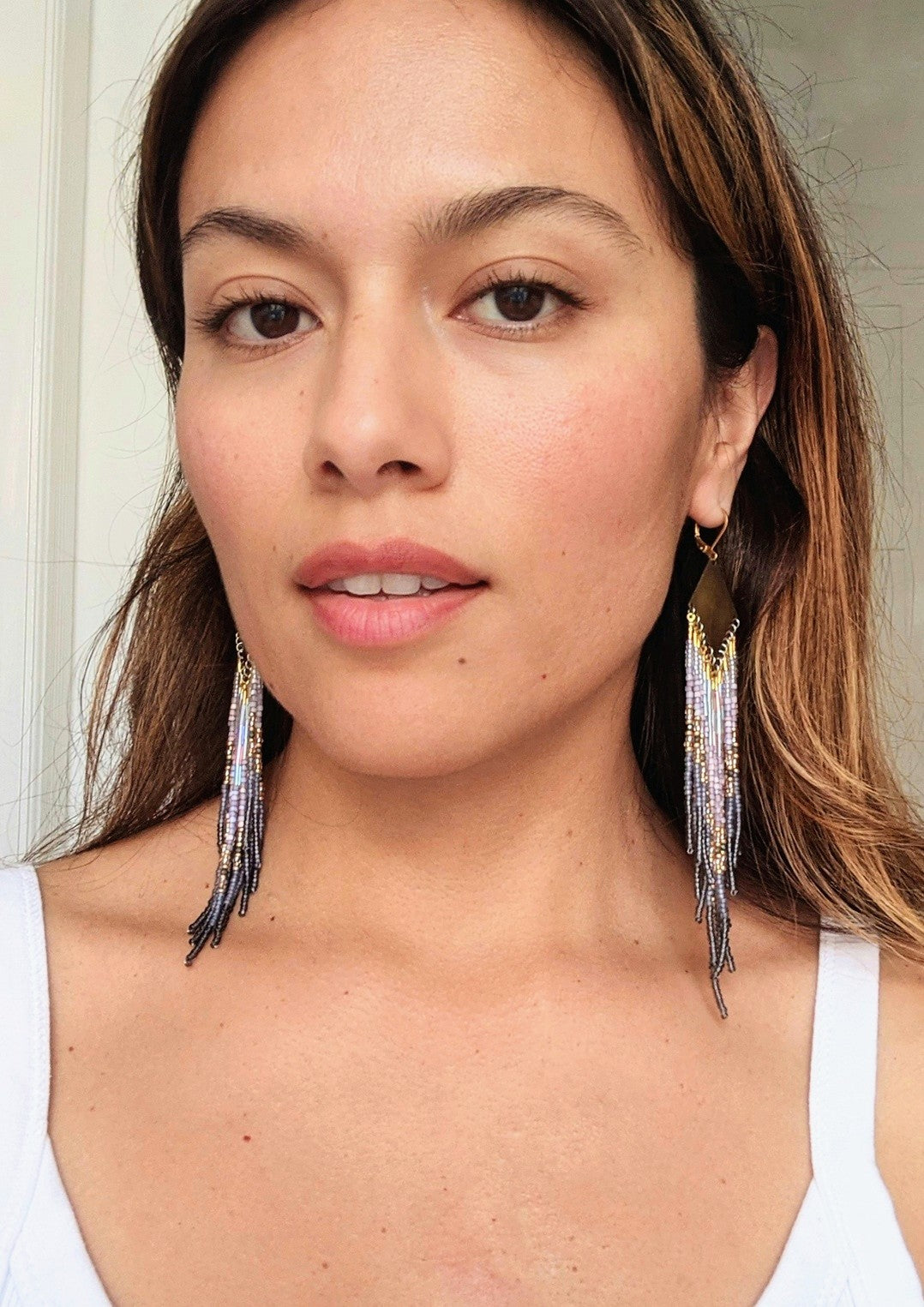 Moon & Milk handcrafted beaded earrings with lavender, gold, grey, and iridescent seed beads on brass diamond-shaped findings, worn by a woman.