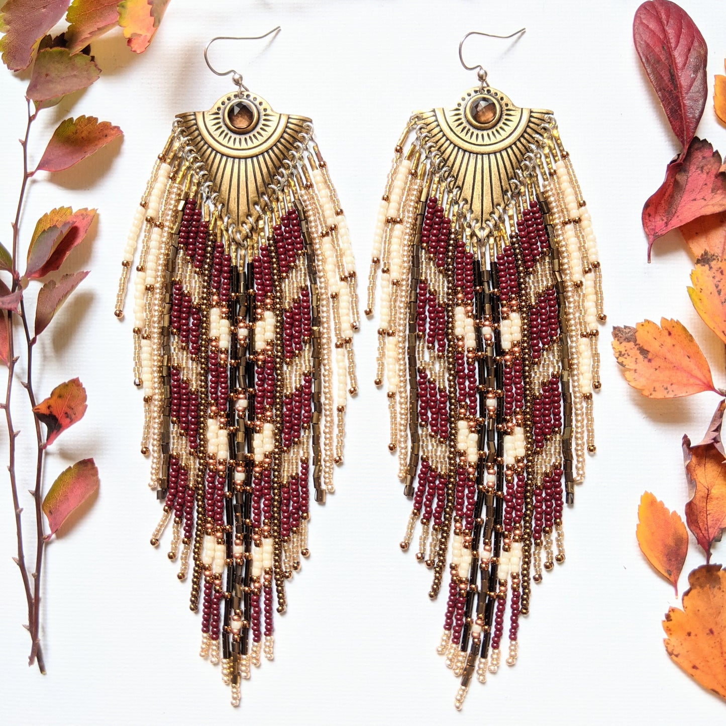 Moon & Milk - Handmade seed bead earrings with maroon, cream, and gold beads, featuring a brass pendant with a central brown stone, displayed with autumn leaves.