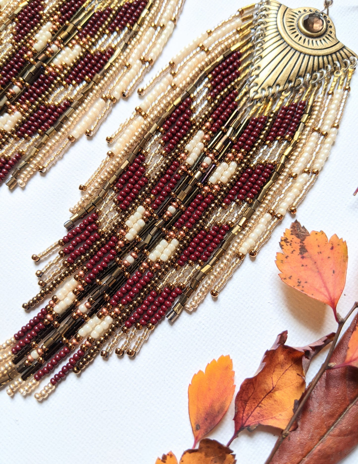 Moon & Milk long beaded earrings with brass fan medallions and cascading burgundy, cream, and gold beadwork, surrounded by autumn leaves on white.