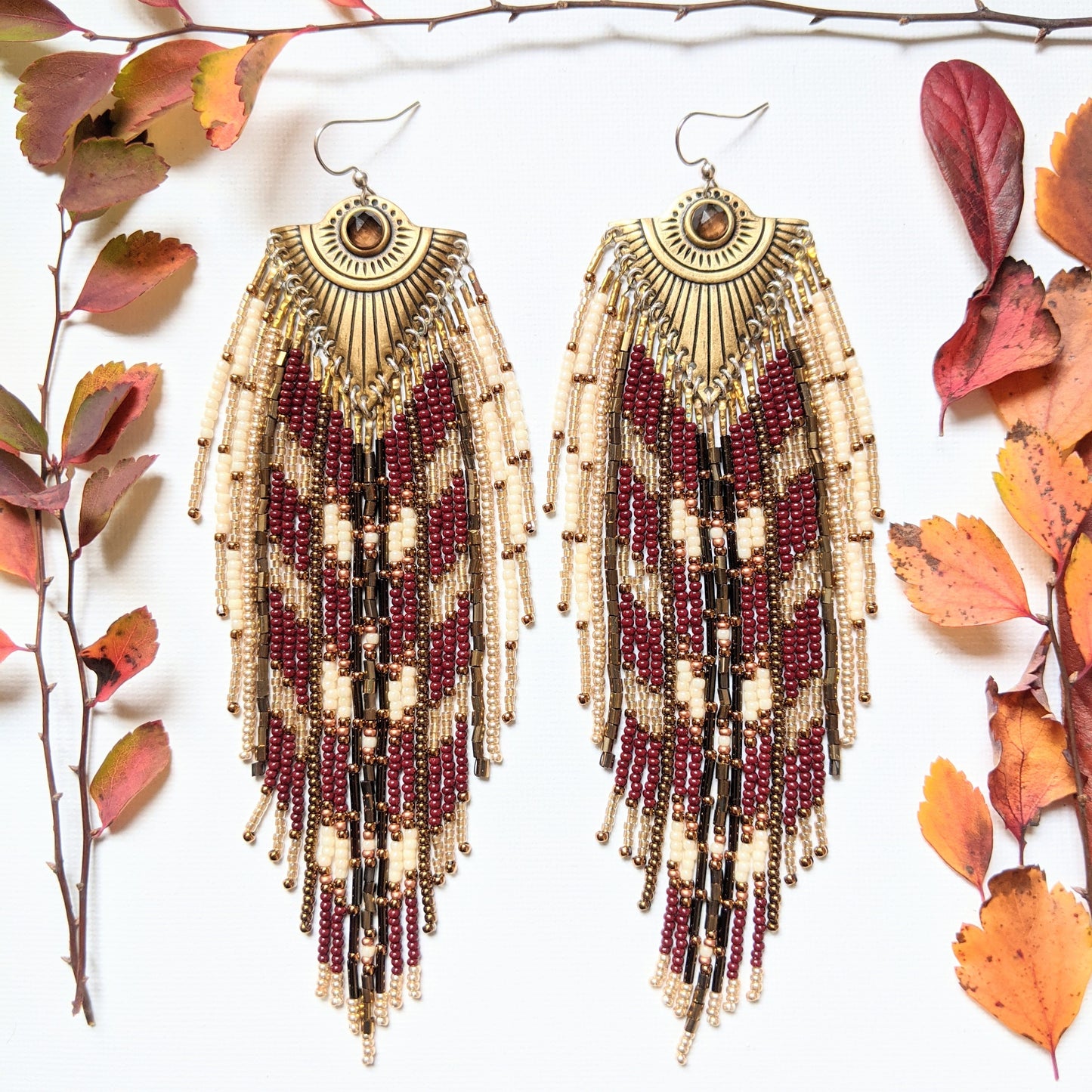 Moon & Milk - Handmade seed bead earrings with maroon, cream, and gold beads, featuring a brass pendant with a central brown stone, displayed with autumn leaves.