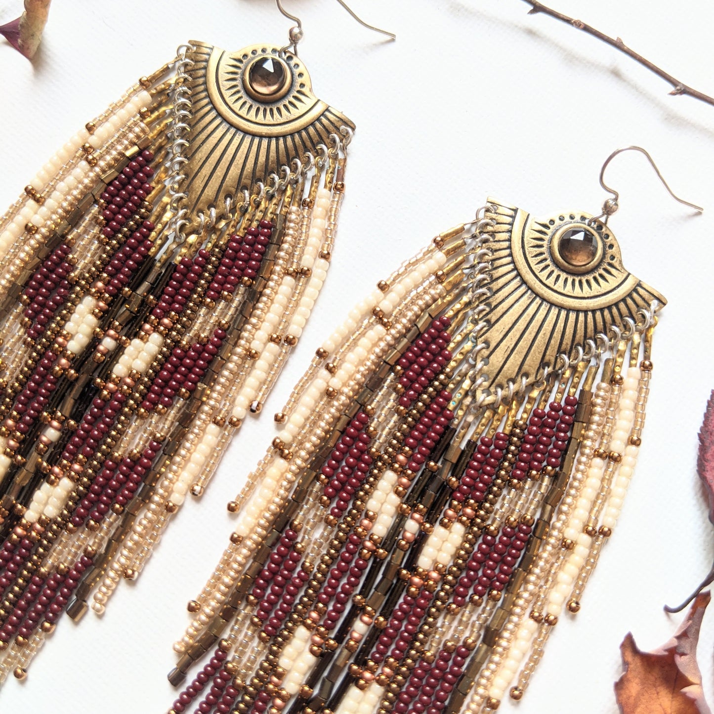 Moon & Milk long beaded earrings with brass fan medallions and cascading burgundy, cream, and gold beadwork, surrounded by autumn leaves on white.