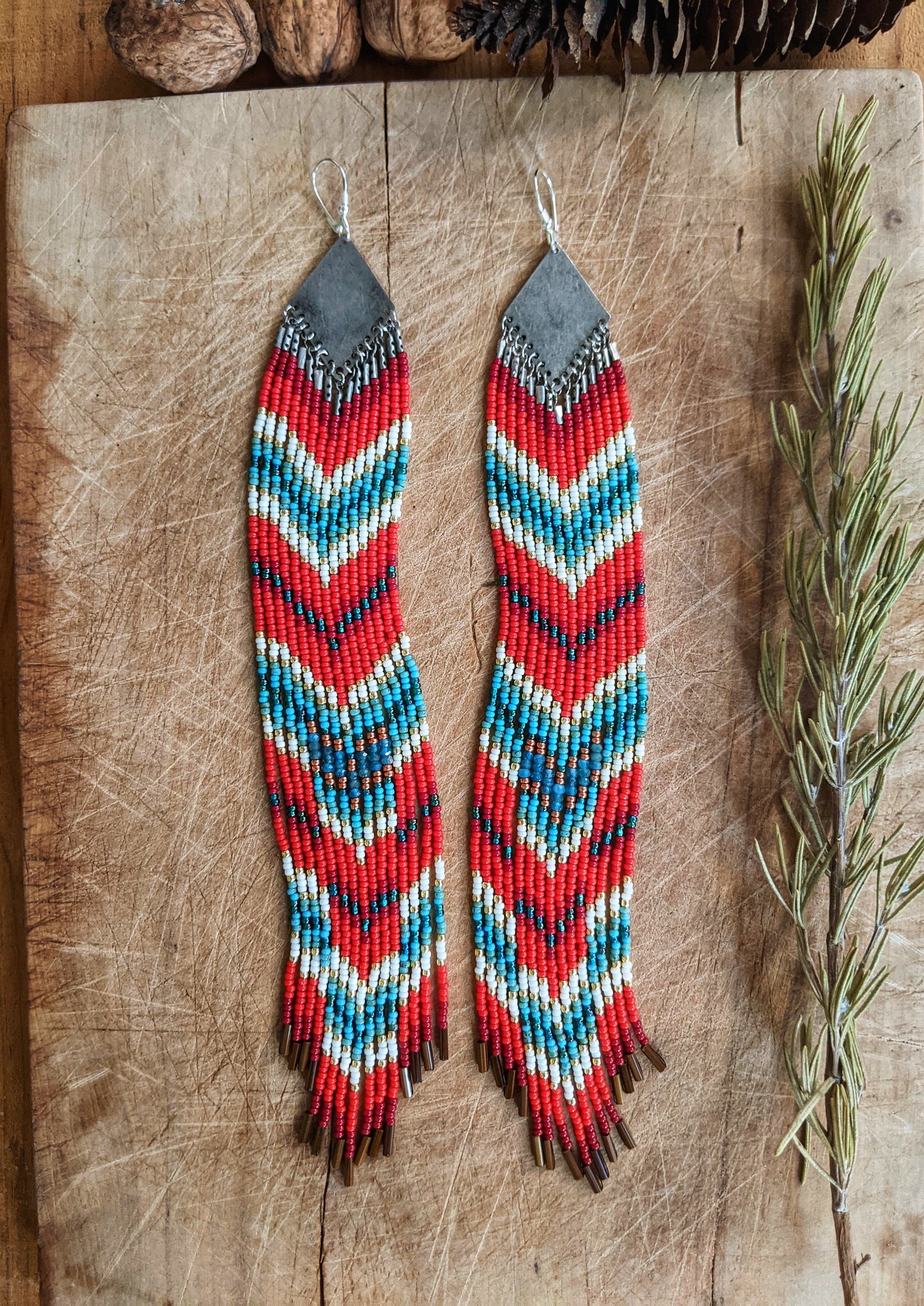 Moon & Milk - Extra long beaded earrings with red, blue, yellow, and white beads in a chevron pattern. Silver diamond-shaped tops. Photographed on a wooden surface with sprigs of rosemary.
