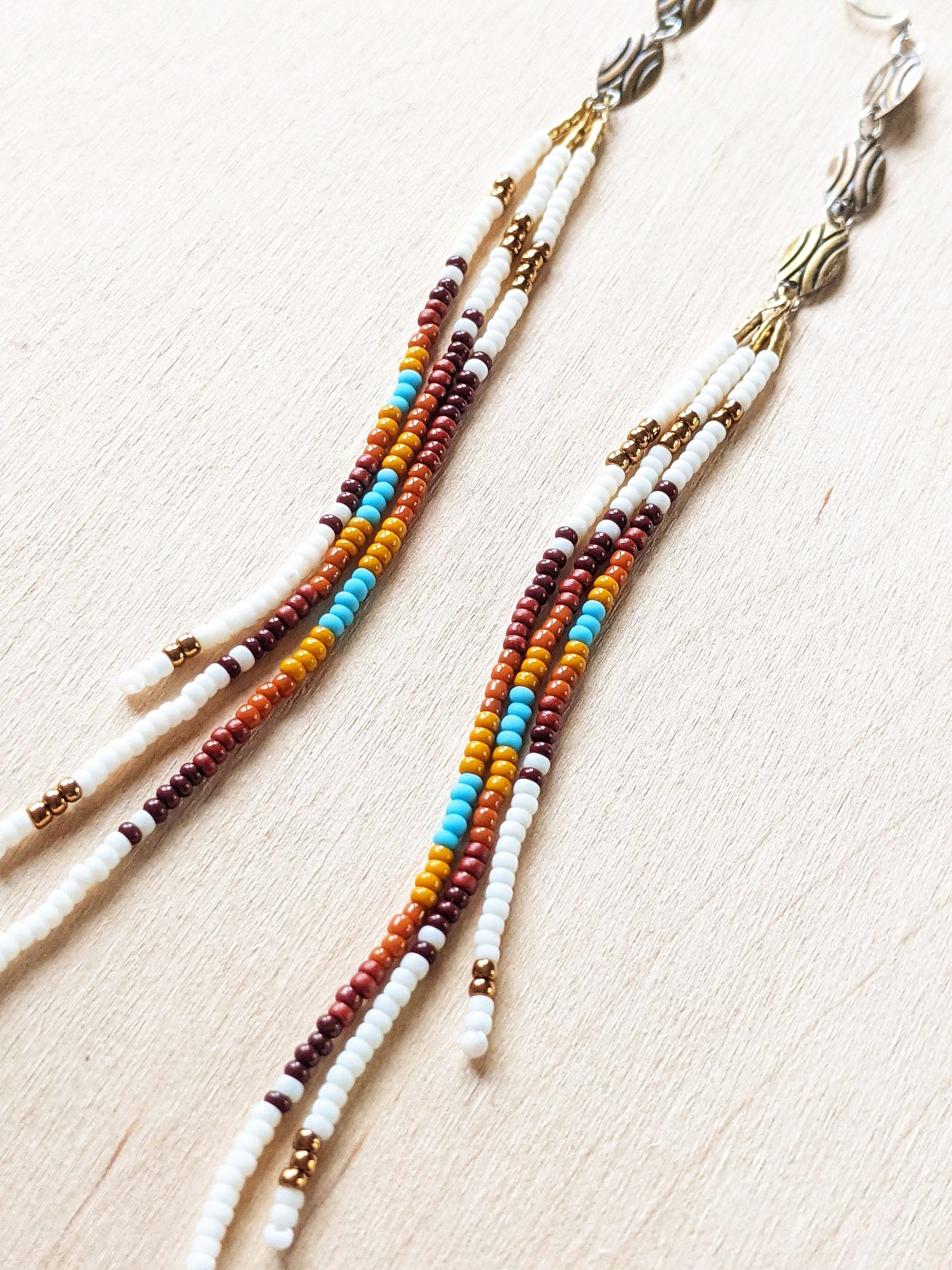 Moon & Milk - Close up of long, multicolored beaded earrings featuring a blend of white, brown, orange, yellow, and turquoise beads, with bronze accents, laid against a light wood background.