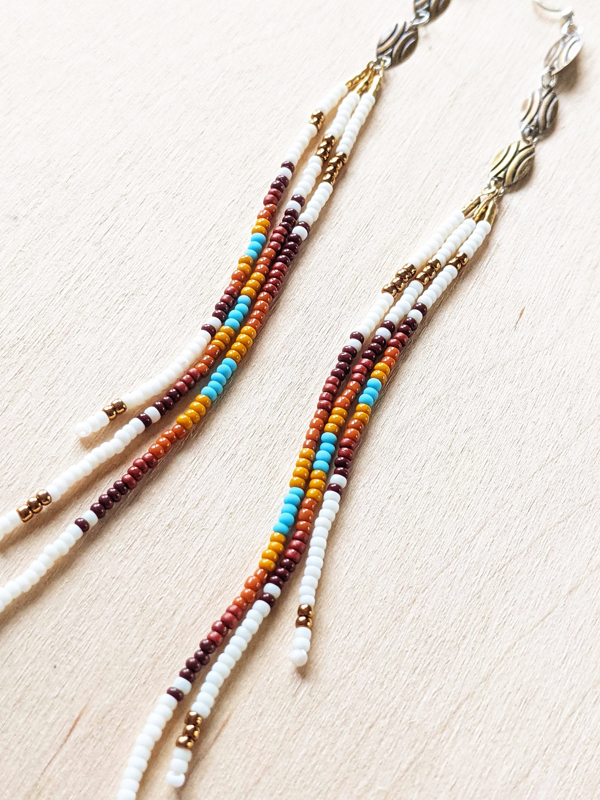 Moon & Milk - Close up of long, multicolored beaded earrings featuring a blend of white, brown, orange, yellow, and turquoise beads, with bronze accents, laid against a light wood background.
