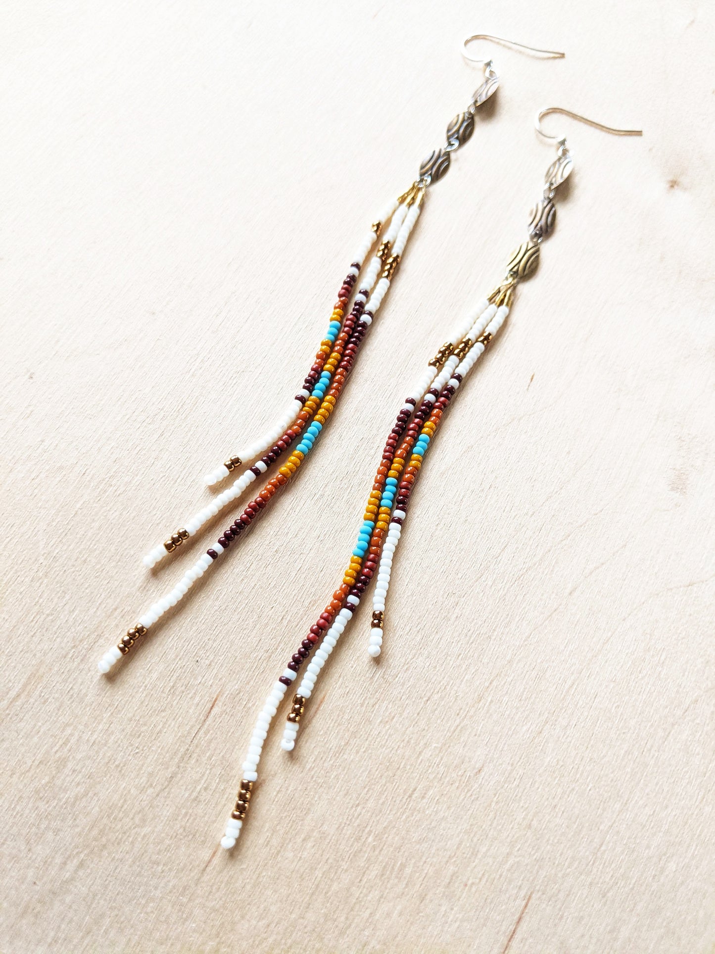 Moon & Milk - Long, multicolored beaded earrings featuring a blend of white, brown, orange, yellow, and turquoise beads, with gold and silver accents, laid against a light wood background.