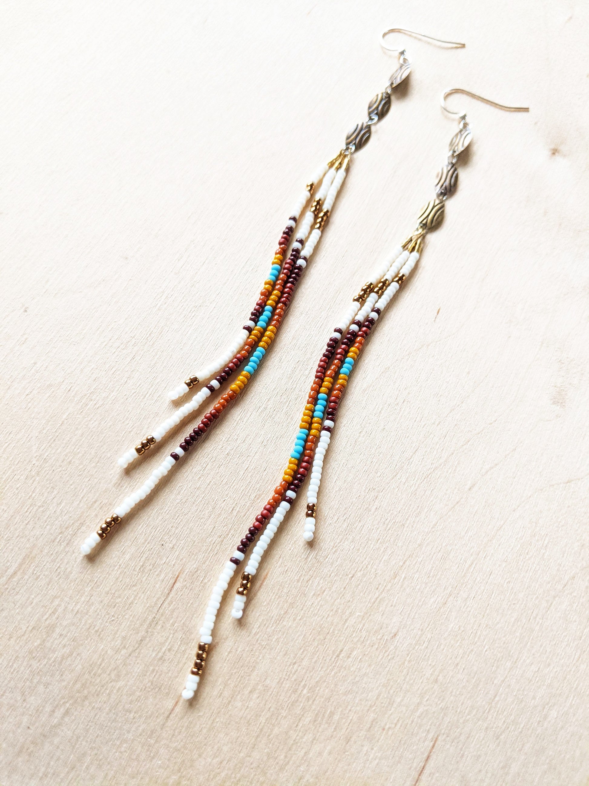 Moon & Milk - Long, multicolored beaded earrings featuring a blend of white, brown, orange, yellow, and turquoise beads, with gold and silver accents, laid against a light wood background.