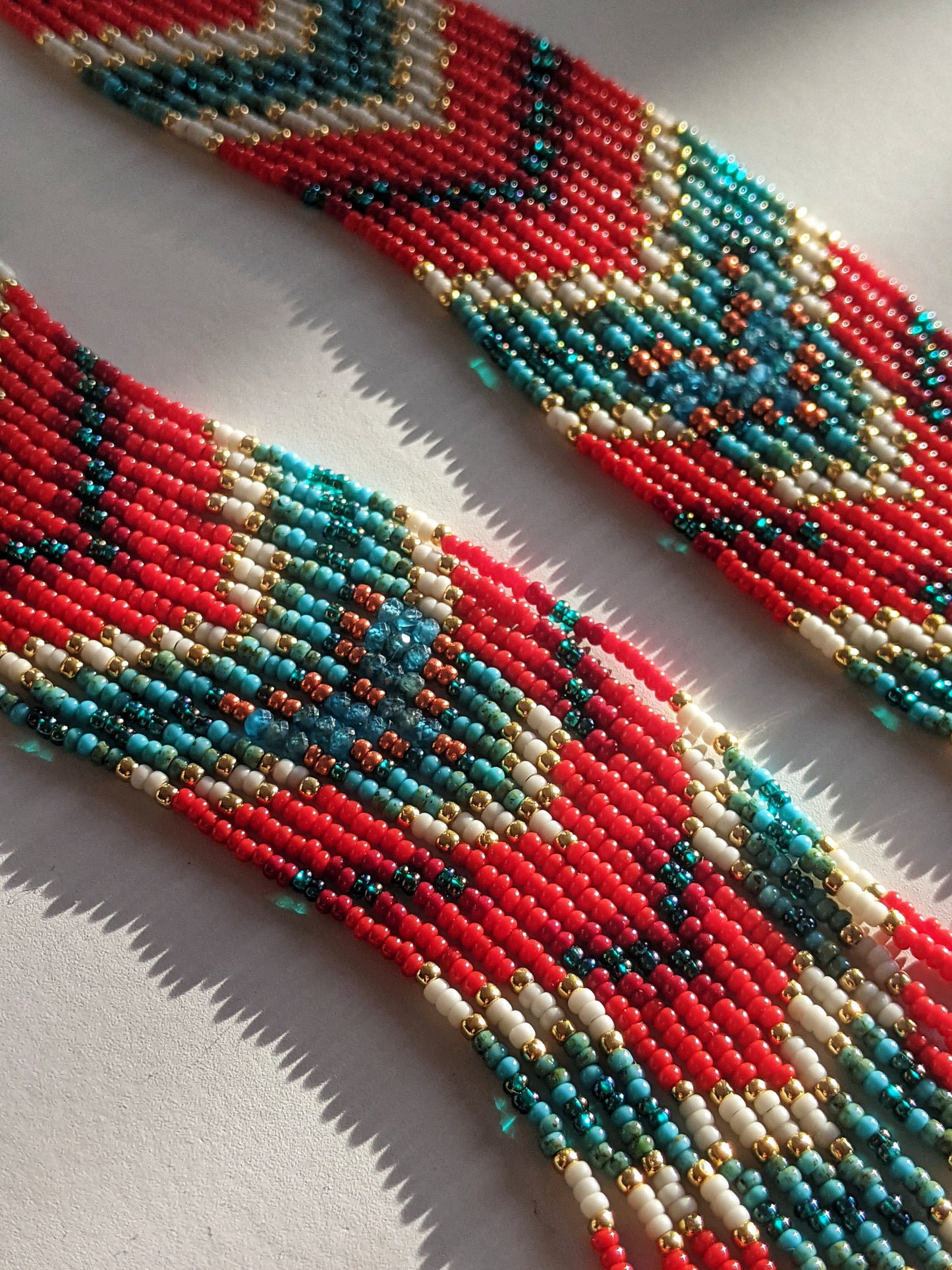 Close-up of beaded earrings in vibrant coral red, turquoise, and gold, featuring a detailed geometric pattern with reflective beads that cast soft shadows. - Moon & Milk