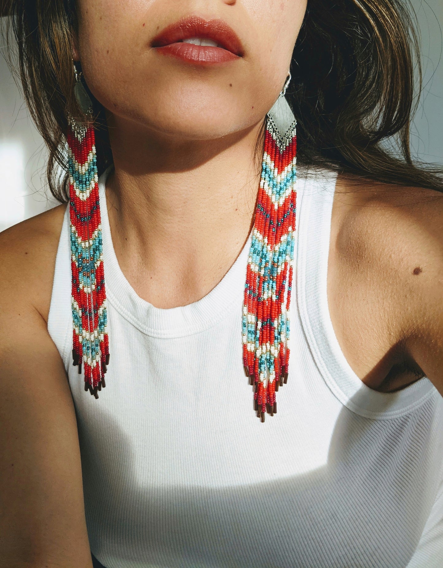 A model wearing long, beaded earrings with red, blue, gold, and white beads in a chevron pattern and silver tops. Styled with a white top. - Moon & Milk
