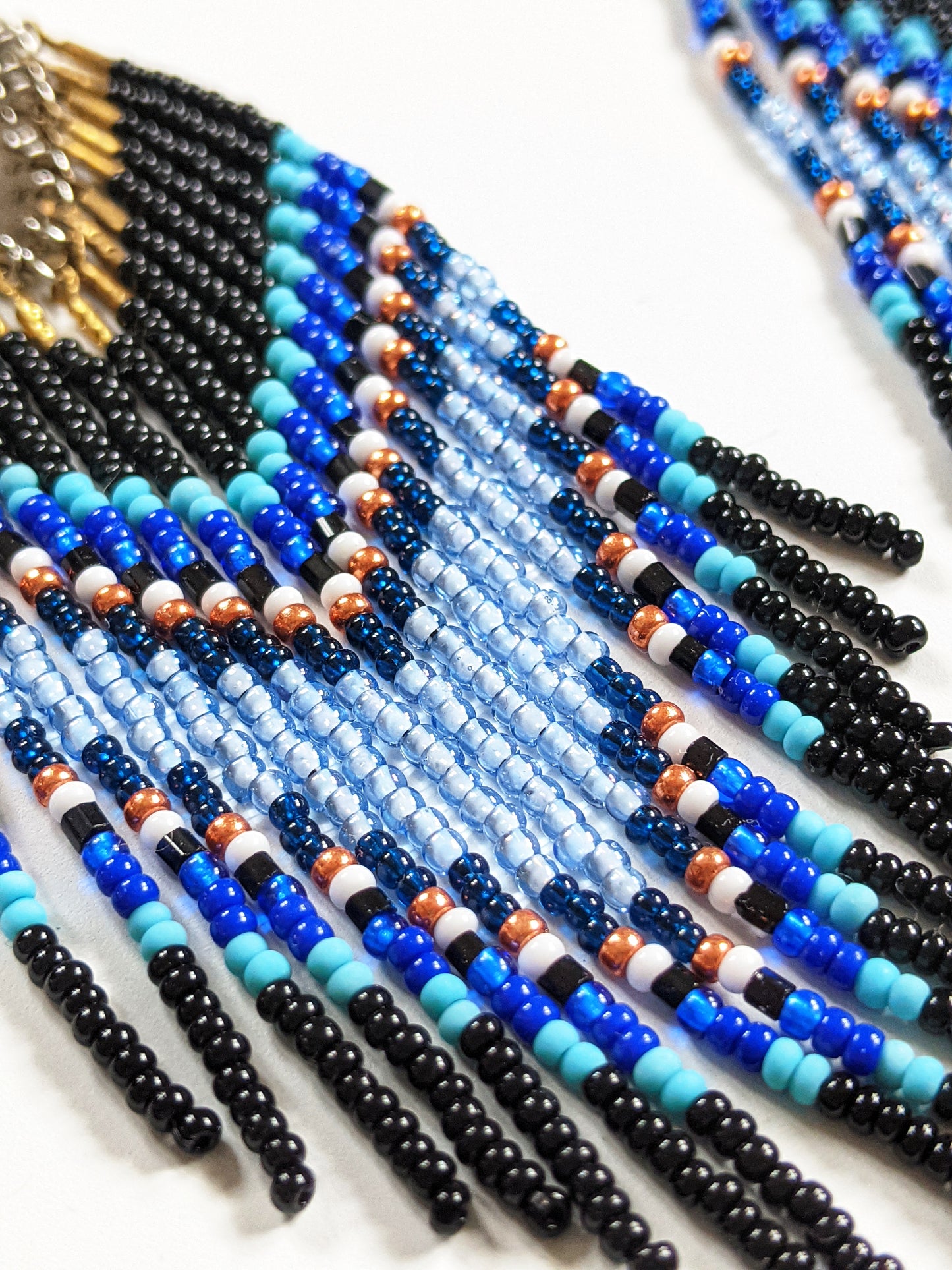 Moon & Milk handmade chevron beaded earrings in black, cobalt, and sky blue with metallic accents, styled on a white plate with fresh flowers.