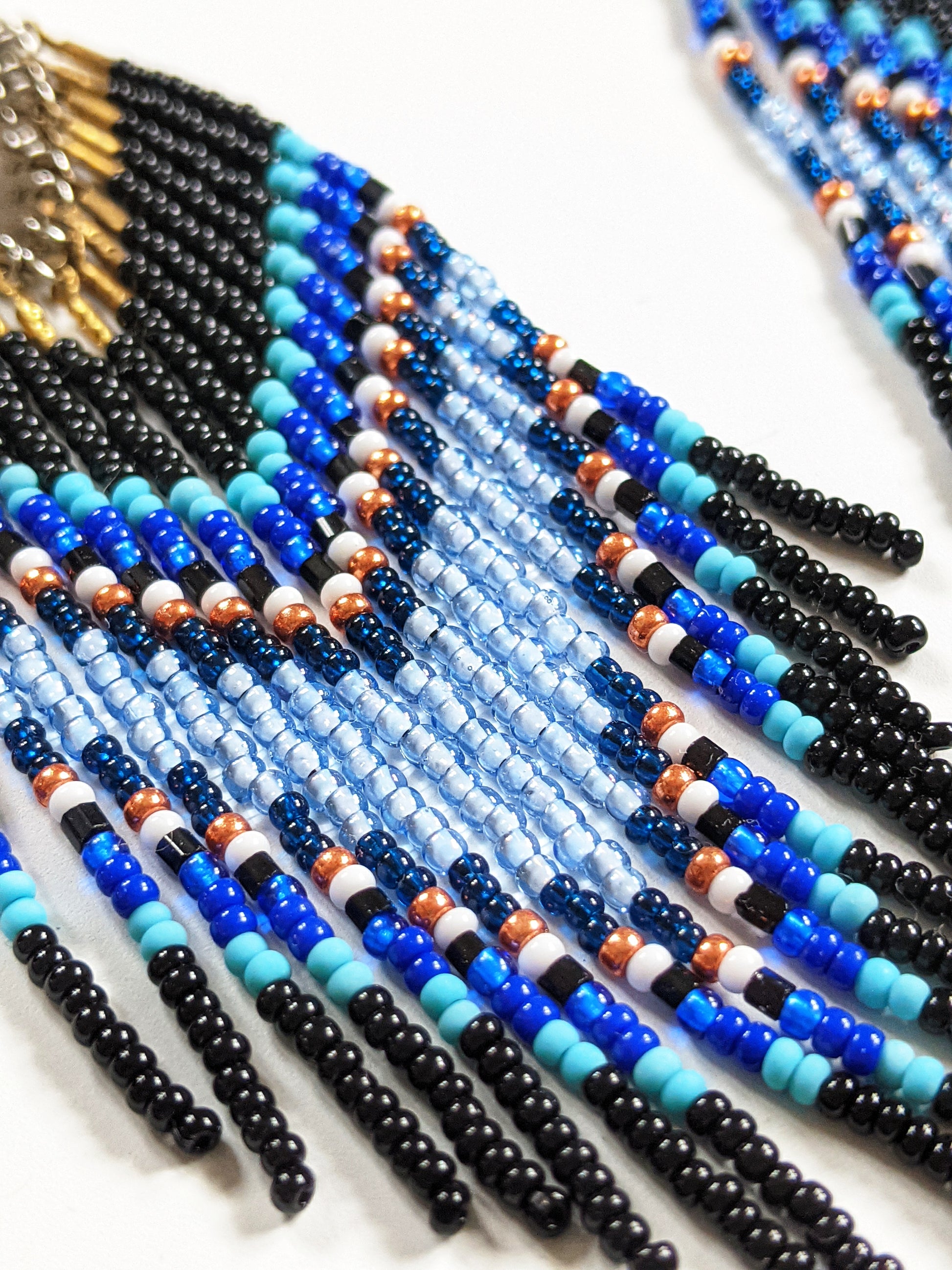 Moon & Milk handmade chevron beaded earrings in black, cobalt, and sky blue with metallic accents, styled on a white plate with fresh flowers.